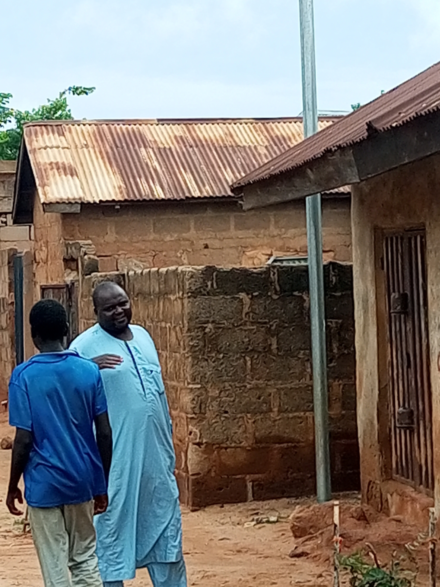 Two males are standing outdoors in an unpaved area. The male on the left is wearing a blue t-shirt and light-colored pants. The male on the right is wearing a light blue, long robe and is facing the other male, with his left arm extended and hand resting on his chest. They are standing in front of buildings constructed of brick and stone, with corrugated metal roofs. A metal pole stands vertically between the two figures. The scene appears to be in Tempure, Nigeria.