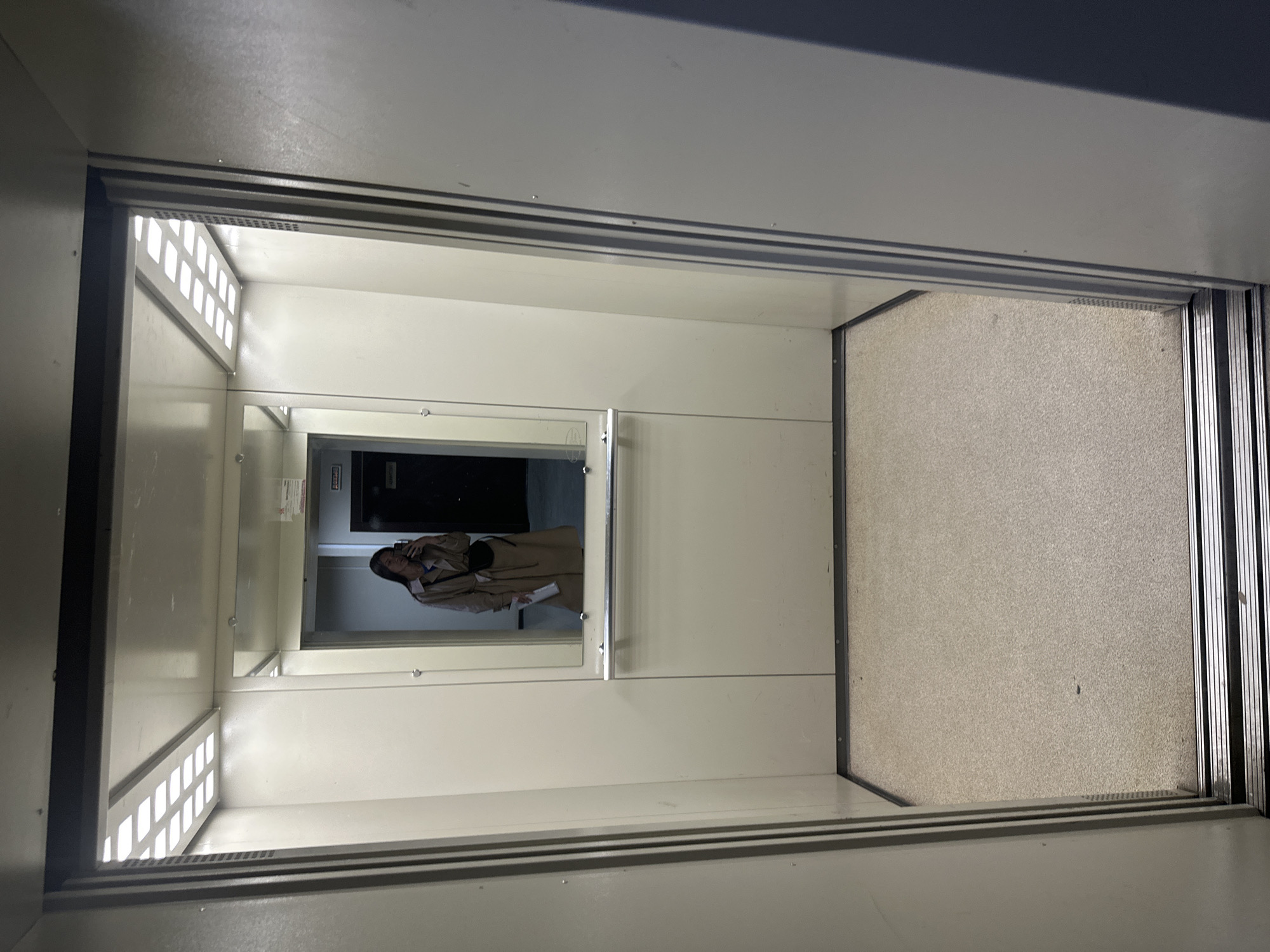 The image shows the interior of a small elevator. The walls are off-white. There is a mirror on one wall, reflecting a person taking the photo. The floor is covered in a textured beige material. There are lights on the ceiling. The location is likely in Izhevsk, Russia, but there is no confirming evidence in the image.