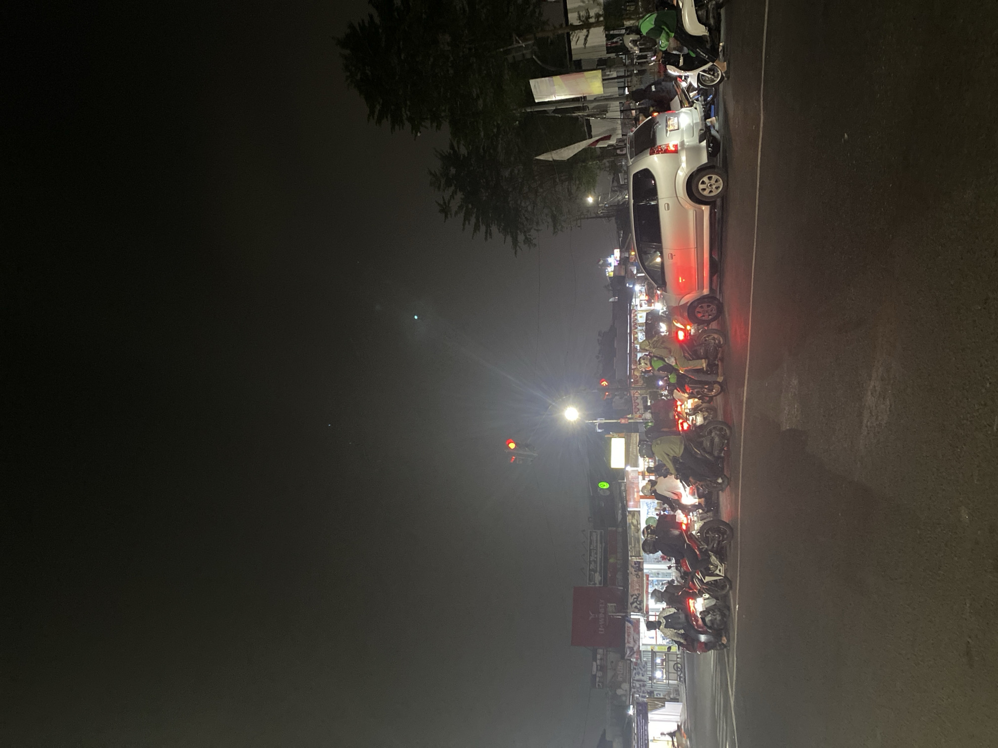 The image depicts a nighttime scene at a traffic intersection in Tangerang, Indonesia, likely during a period of traffic congestion. The foreground shows a road with multiple motorcycles and one silver minivan stopped at the intersection. Several individuals are riding the motorcycles, some wearing helmets. The minivan has its brake lights illuminated. In the background, there are buildings with illuminated signs, suggesting commercial establishments. Traffic lights are visible, with one appearing red and another green. Trees are present on the right side of the image. The sky is dark and overcast.