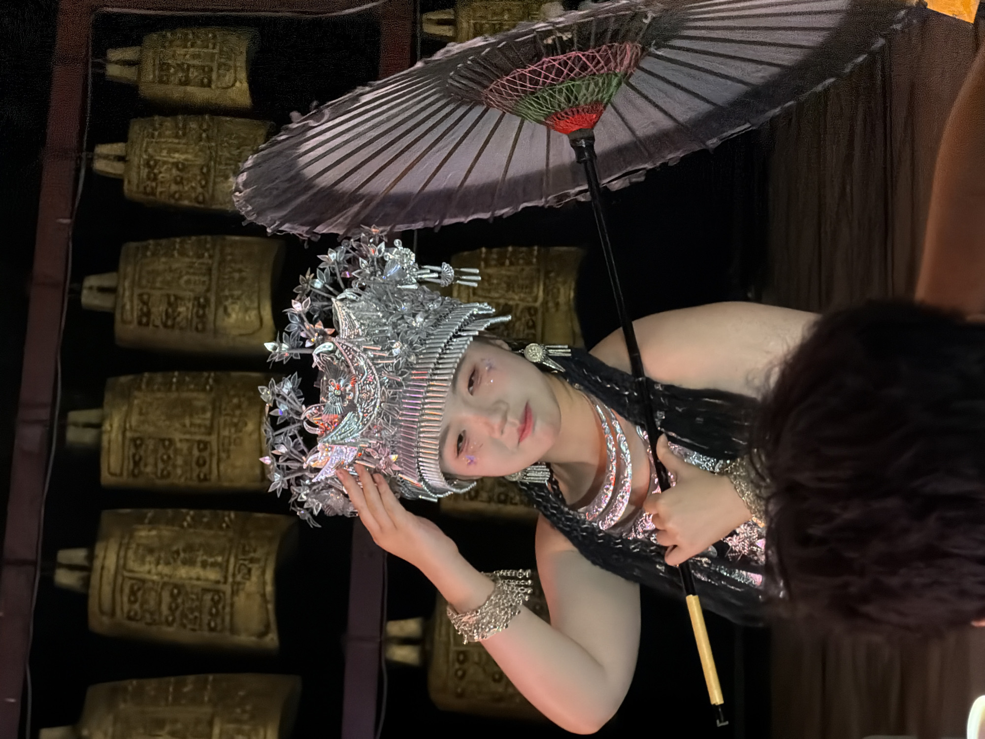 In Yuzhong District, China, a person adorned in elaborate traditional attire, likely a costume, poses for a photograph. They wear a striking silver headdress embellished with intricate designs and sparkling accents. A silver necklace and bracelets complement the headwear, adding to the regal appearance. The individual holds a delicate, dark umbrella, its fabric patterned and its stem a golden yellow. Behind them, a series of large, golden cylindrical objects are arranged vertically, each featuring embossed designs that suggest historical or cultural significance. The lighting is dim, with spotlights illuminating the subject and creating a dramatic effect. The overall scene evokes a sense of performance or ceremonial display, possibly at an indoor cultural event or exhibition. The individual's expression is gentle and perhaps slightly reserved, adding to the dignified presentation.