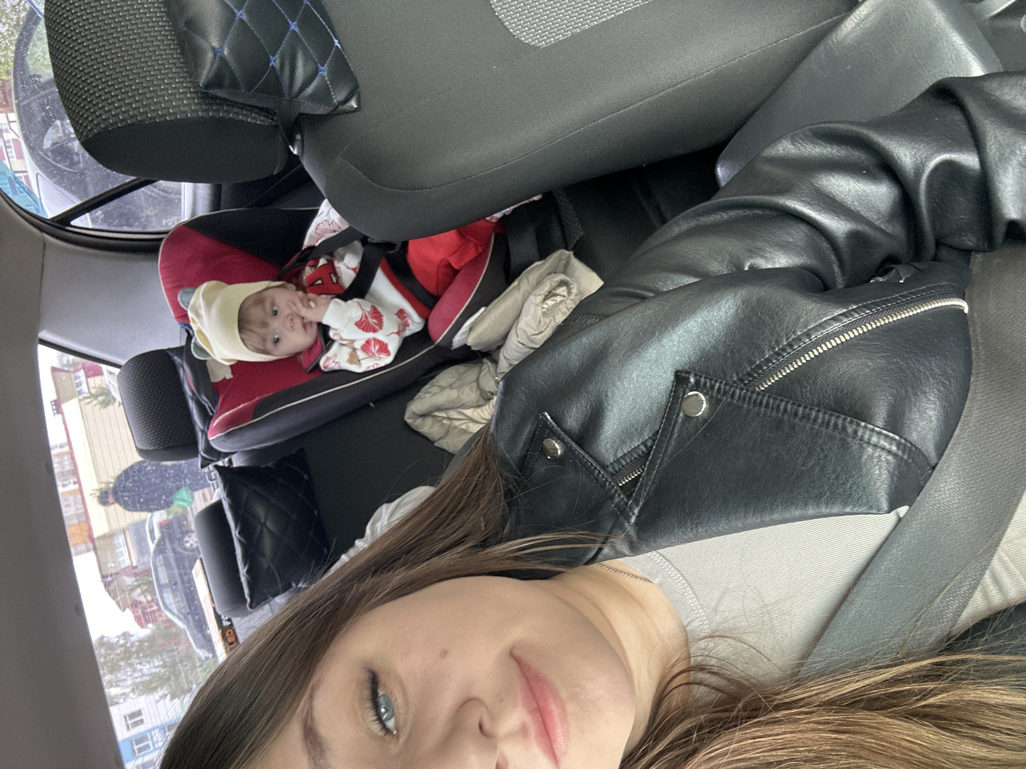 The image shows a woman taking a selfie inside a car. In the background, a baby is visible in a car seat. The location is Chaikovsky, Russia, as determined by metadata not visible in the image but available to the AI. The woman is wearing a black leather jacket and a seatbelt. The car interior includes grey seats. Outside the window, buildings and other cars are visible. The baby is wearing a light-colored hat and a patterned outfit.