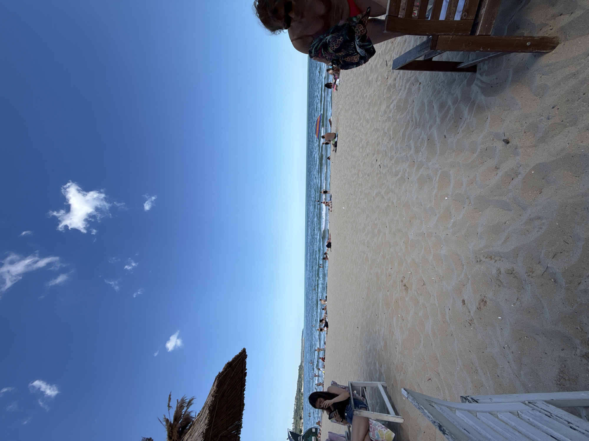 This is a photograph taken on a beach in Varna, Bulgaria, on a sunny day with a clear blue sky and a few scattered clouds. The foreground is dominated by sand with some small ripples, and two chairs are visible. To the left is a white, weathered wooden chair with a woman sitting in it, looking at her phone. To the right is a brown wooden chair. In the midground, several people are scattered on the sand and in the sea, enjoying the water. Some are lying down, others are sitting, and some are wading or swimming. Beach umbrellas are visible, including a colorful one in the center. The sea is calm with gentle waves. A thatched umbrella is partially visible in the upper left corner, casting a shadow on the sand. In the far background, a treeline and some buildings can be seen along the coastline. The person on the right side of the image is partially visible, appearing to be a woman in a red swimsuit, holding a patterned cloth.