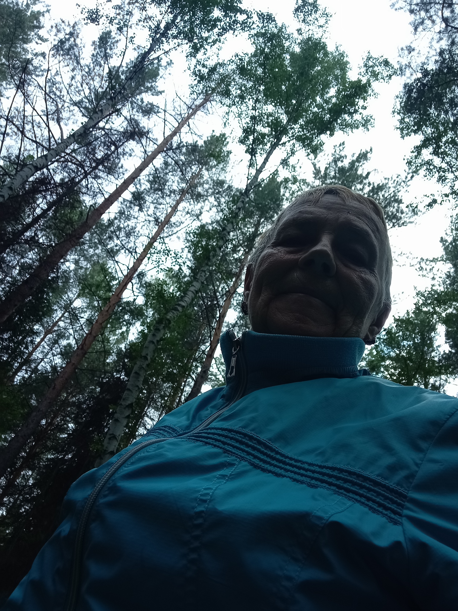 A low-angle, outdoor, close-up shot features a person in a teal jacket against a background of tall trees reaching towards an overcast sky. The person's face, partially obscured by shadow, is looking up and slightly to the left. The jacket is zipped up to the neck, and textured detailing is visible across the chest. The environment suggests a forest or wooded area. This image is consistent with being taken in the city of Degtyarsk, Russia, where dense forests are common.
