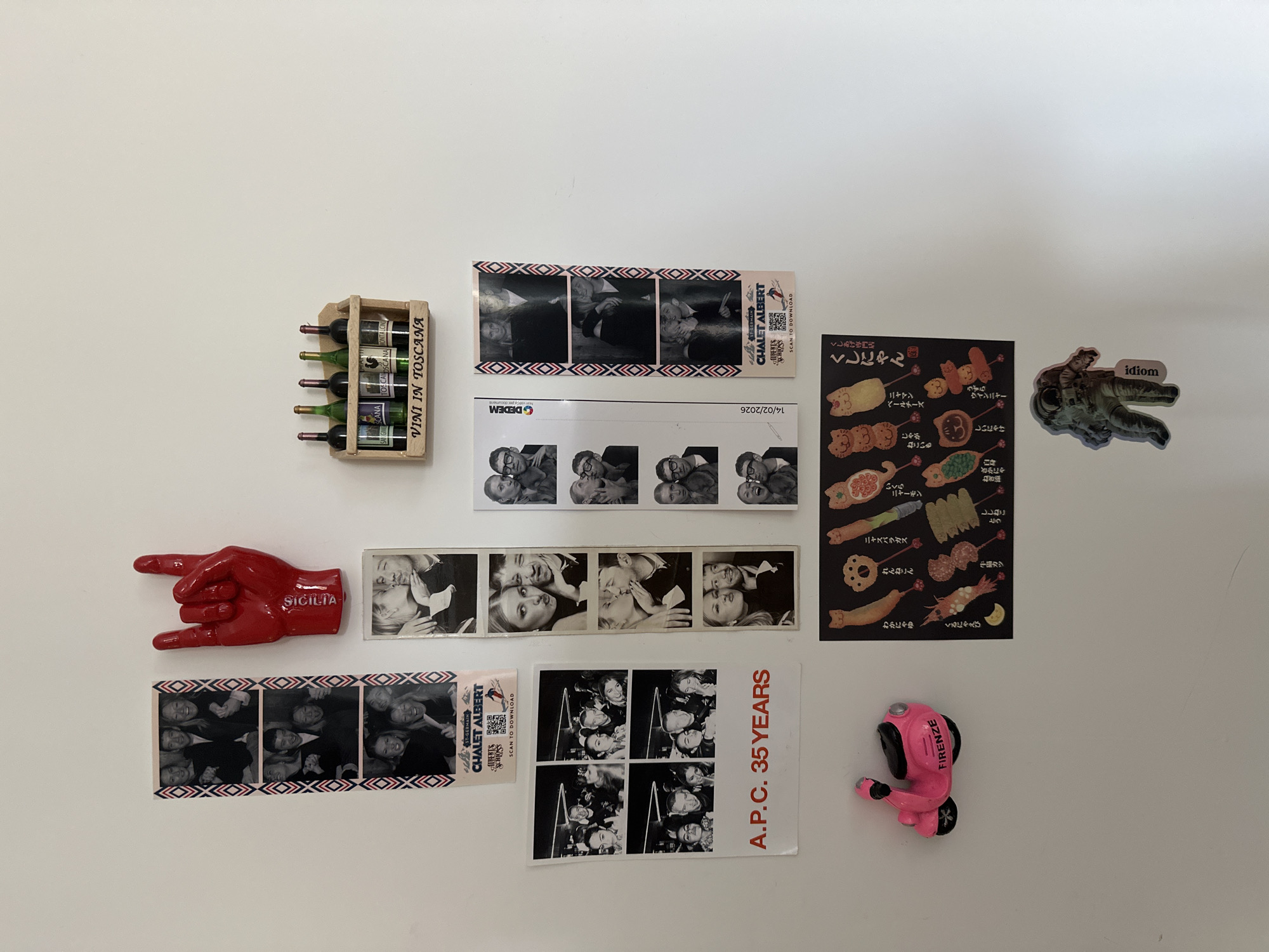 On a white background, presumably a wall, are several objects. These include a miniature wooden wine rack containing five bottles of wine labeled "VINI IN TOSCANA" and "TOSCANA." There is also a red hand-shaped magnet labeled "SICILIA." Several photo booth strips are also present, capturing black and white images of people. One of these strips is branded "CHALET ALBERT" and another has a "DEDEM" logo with a date "14/02/2026." There is a small menu or advertisement for "くしにゃん", a Japanese restaurant serving fried skewered food. An astronaut sticker with the word "idiom" is also included. Finally, there is an "A.P.C. 35 YEARS" advertisement and a pink hairdryer-shaped magnet labeled "FIRENZE." The image location is not in London, but includes magnets and photos from Italy, a Japanese restaurant advertisement, and French fashion advertisement.