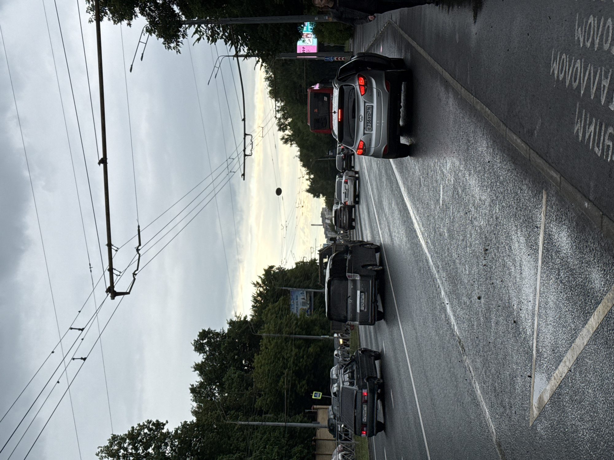 A street scene in Kazan, Russia, on a cloudy day. The wet asphalt road is lined with trees and overhead trolley wires. Several cars are driving on the road, including a dark hatchback, a grey minivan, and a silver SUV. A person is walking on the sidewalk on the right side of the image. There are also some billboards visible in the background.