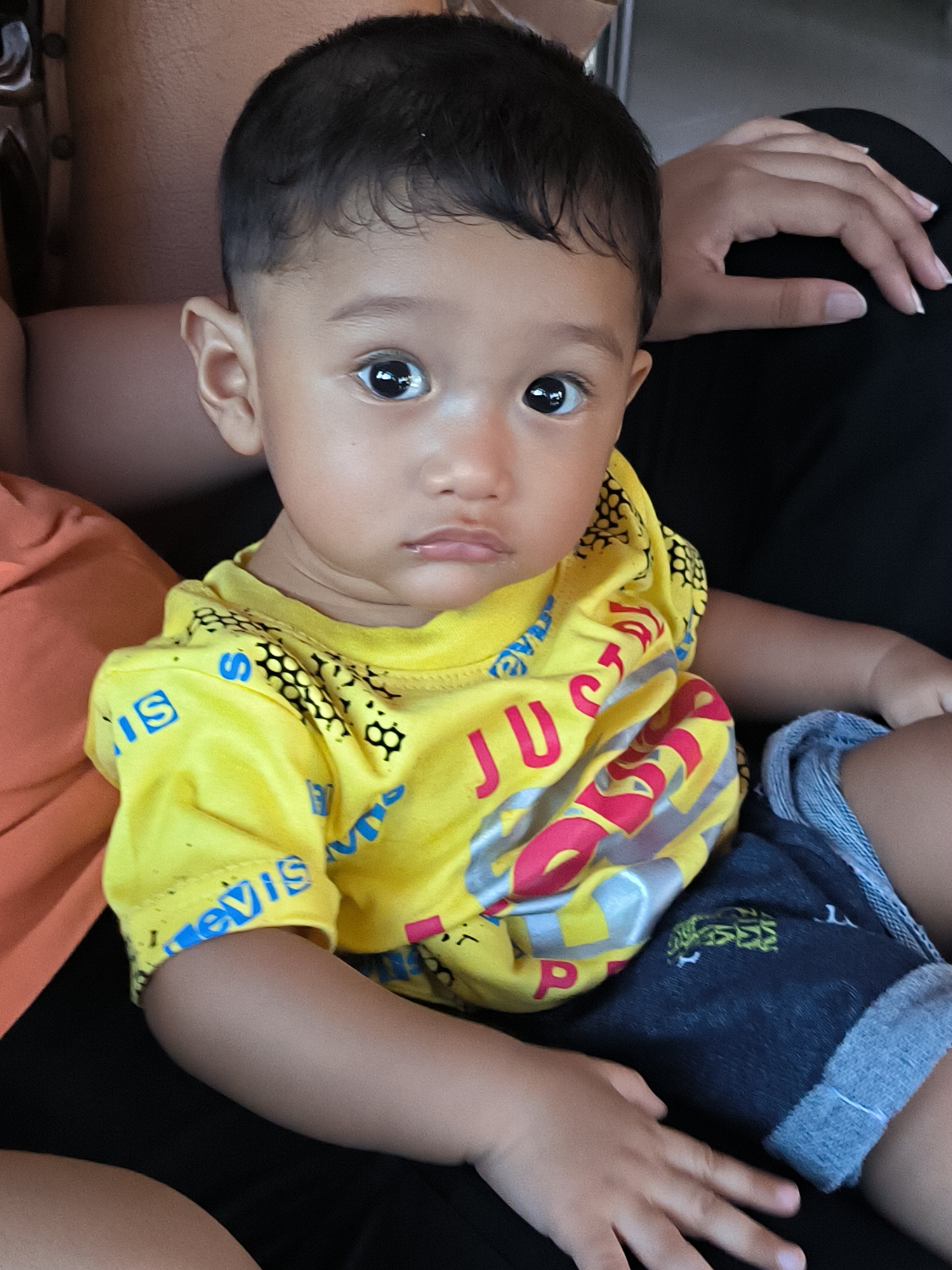 A close-up shot depicts a young child seated in an unknown location, possibly in the city of Merbopuntong, Indonesia. The child is wearing a yellow t-shirt with lettering and blue denim shorts with rolled cuffs. The child is being held by an adult, with partial views of their skin and clothing visible on either side of the child. The adult has their hand resting on the child's right side. The environment is not clearly defined, providing limited context to the scene.