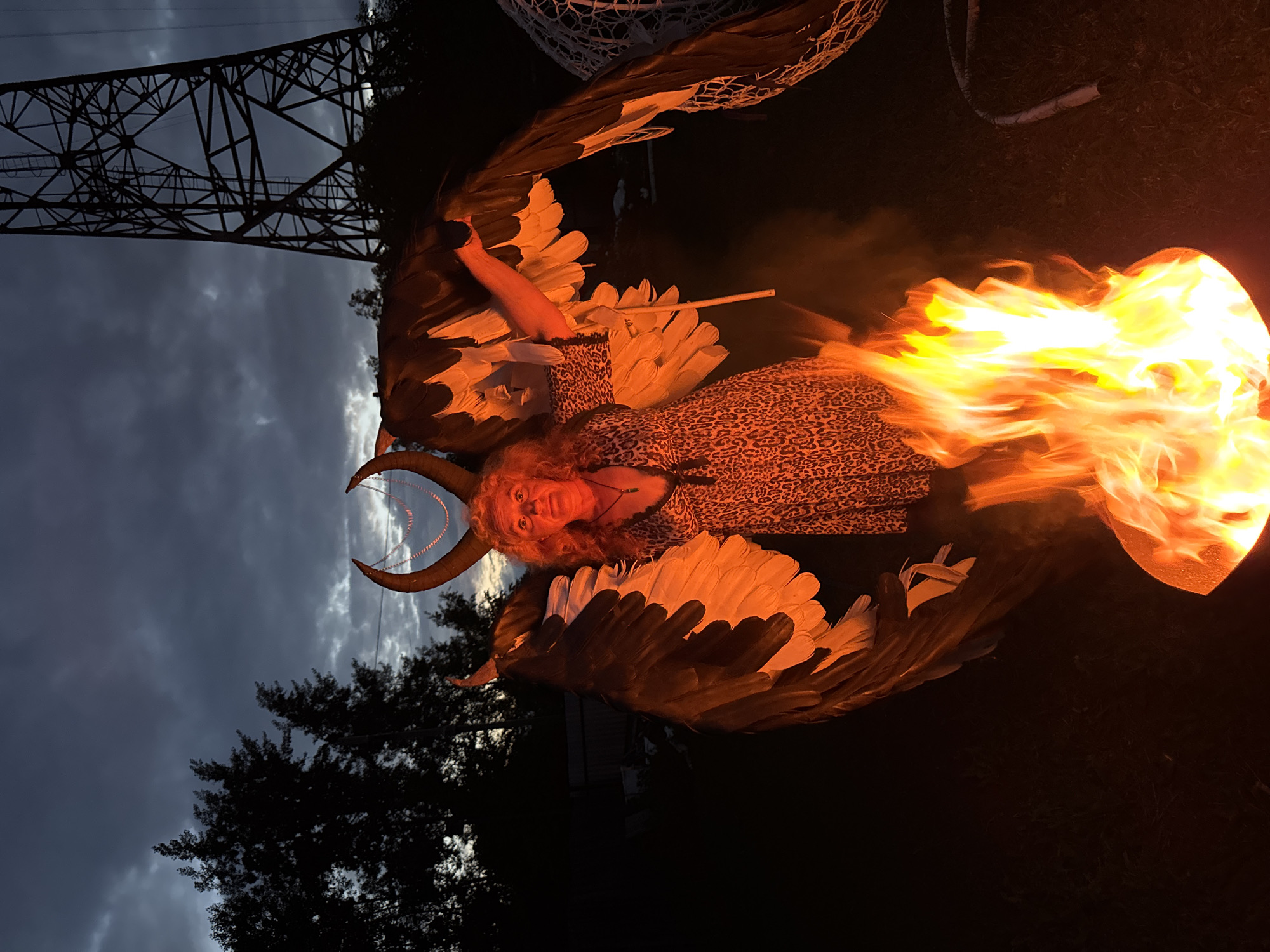A woman with large, dark feathered wings and horns stands in front of a blazing fire pit at dusk. She is wearing a leopard-print dress and a necklace. The background features trees, a power line tower, and a cloudy sky. The scene appears to be in a backyard in the city of Chaikovsky, Russia.