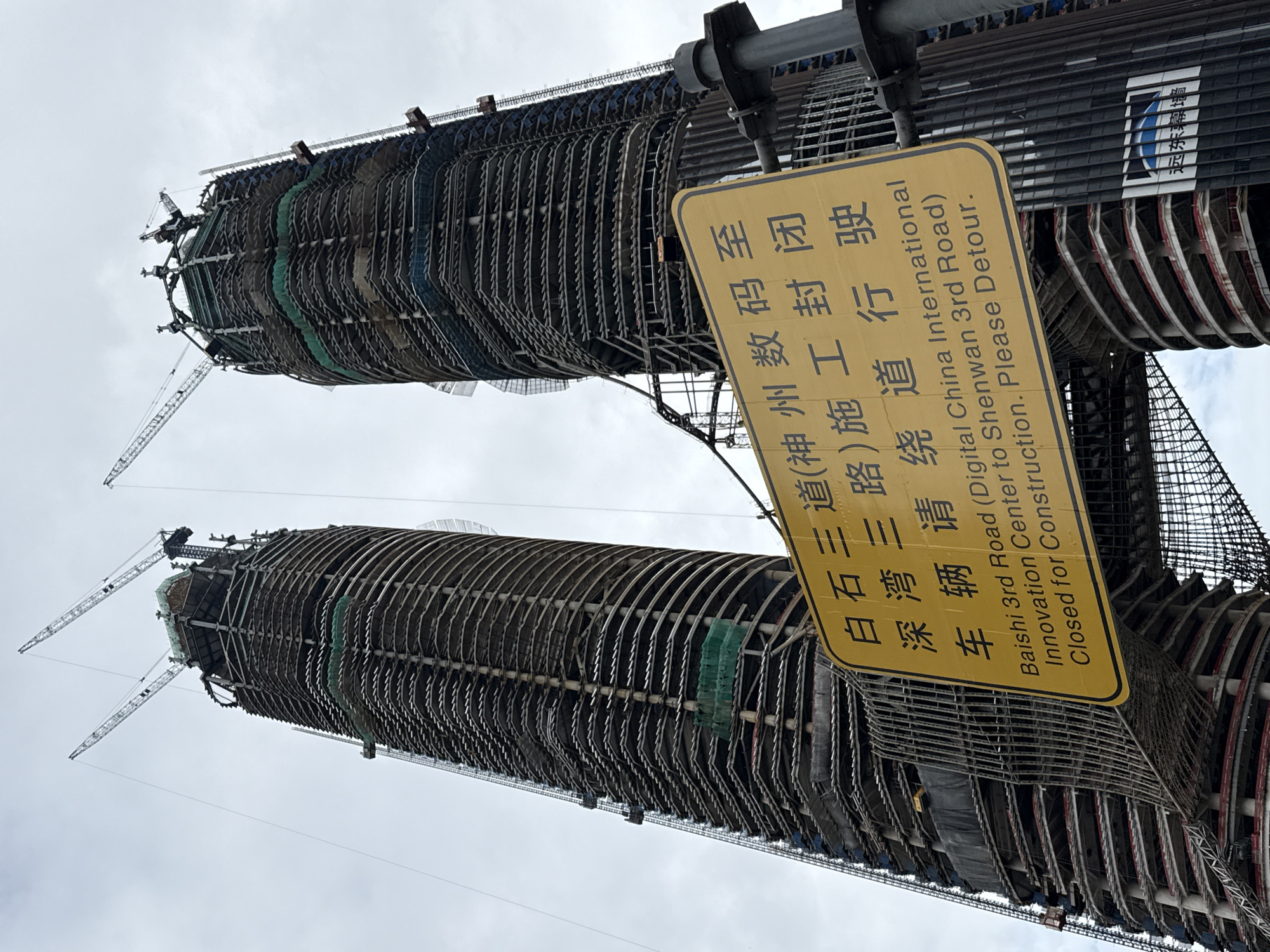 Two massive, unfinished skyscrapers, their skeletal framework exposed, dominate the frame against a cloudy sky. Multiple construction cranes are visible, extending from the tops of the towers and to the side, indicating ongoing work. A bright yellow, diamond-shaped sign stands prominently in the foreground, displaying bilingual text in Chinese and English. The Chinese text reads "白石三道(神州数码至深湾三路)施工封闭", and the English text beneath it states, "Baishi 3rd Road (Digital China International Innovation Center to Shenwan 3rd Road) Closed for Construction. Please Detour." The sign is attached to a metal pole, and the base of one of the skyscrapers is visible behind it, showing concrete floors and metal reinforcements. The overall impression is a scene of large-scale urban development and infrastructure work in progress. The location is identified as Nanshan District, China.