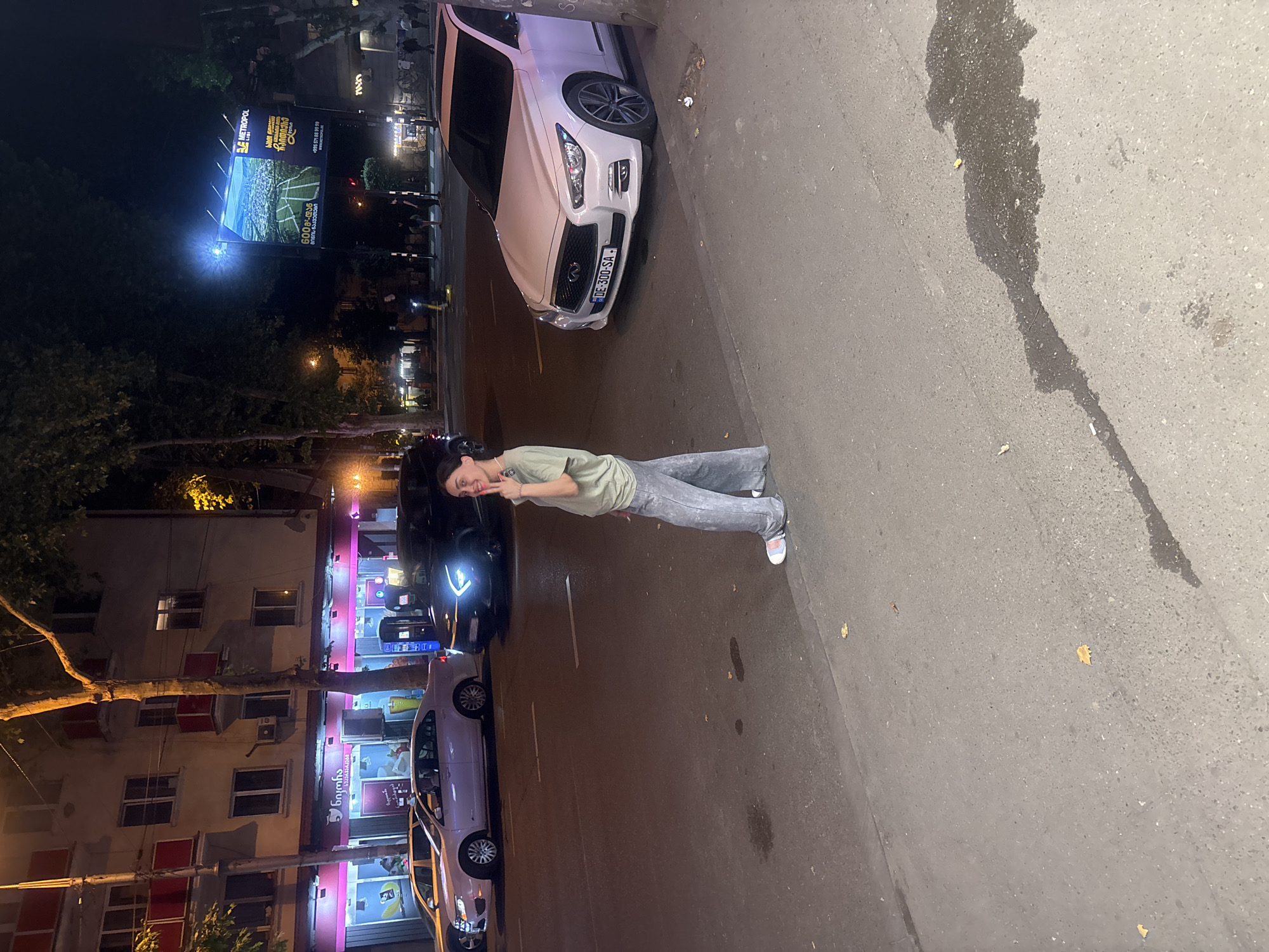 A young woman stands on a sidewalk at night, facing a street in Tbilisi, Georgia. She is wearing a light green t-shirt and baggy gray pants, and she is holding up two fingers to the camera. Behind her, cars are parked and driving on the street, and a building with illuminated storefronts is visible. In the background, a large billboard advertising "Metropol" is lit up. The street is wet, suggesting it may have recently rained. The overall atmosphere is urban and nighttime.
