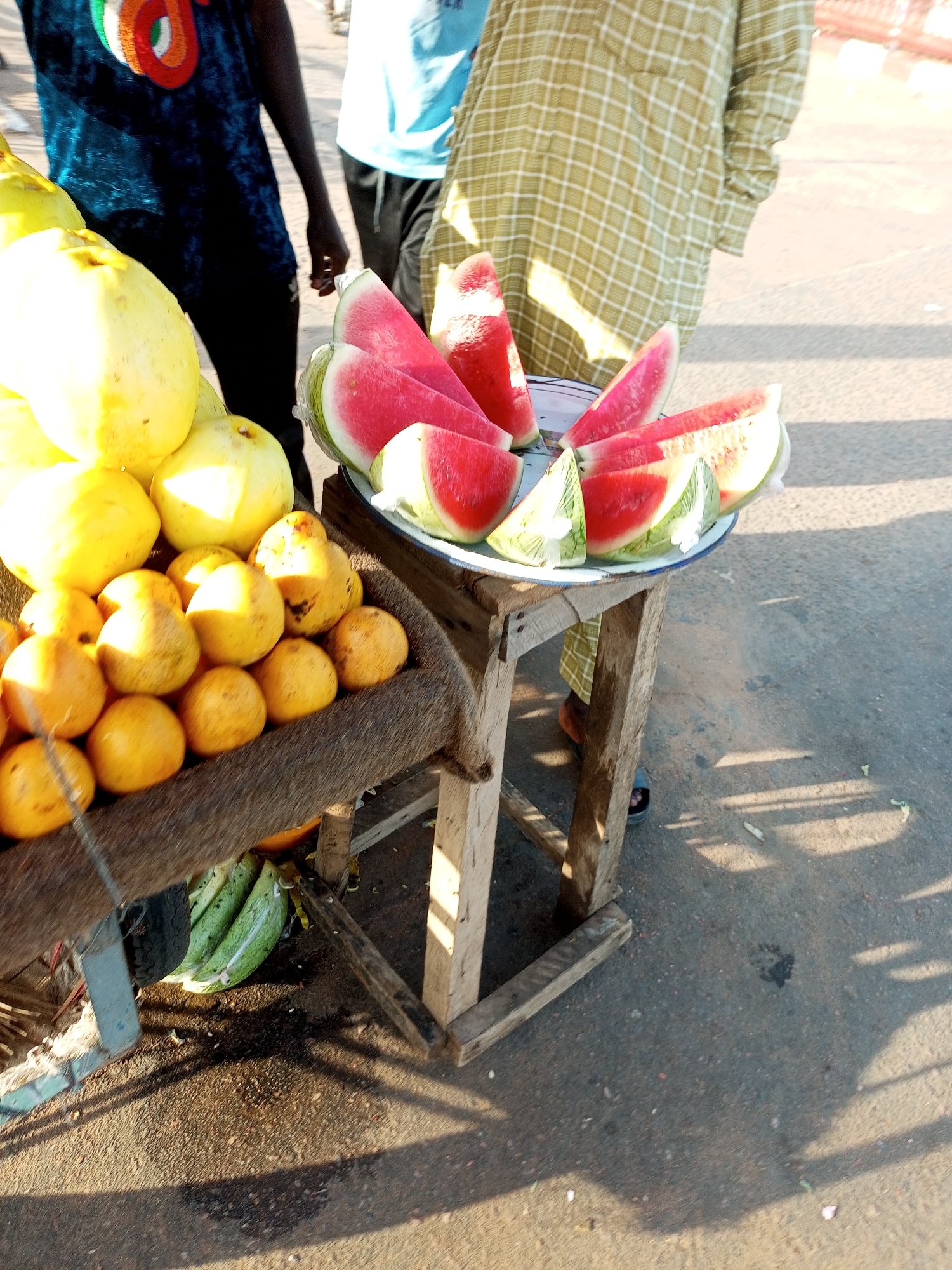 The image depicts a street-side fruit stall in Tumfure, Nigeria. In the foreground, a variety of fruits are displayed. A wooden crate, lined with a furry material, holds an assortment of small, round, orange-colored fruits, possibly tangerines or small oranges. Above this, larger, pale yellow fruits, likely guavas, are piled high. To the right, a metal tray holds several slices of watermelon arranged in a fanned pattern.
Two individuals are partially visible in the background. One, wearing a dark blue t-shirt with a circular design, stands to the left. To the right, another person is seen wearing a long, light green garment with a checkered pattern. They are standing near the fruit stall, which is supported by rough, wooden legs. The ground is paved asphalt, with shadows cast from the stall and the surrounding environment. In the lower left of the image, several long, green fruits, resembling plantains or large bananas, are visible.