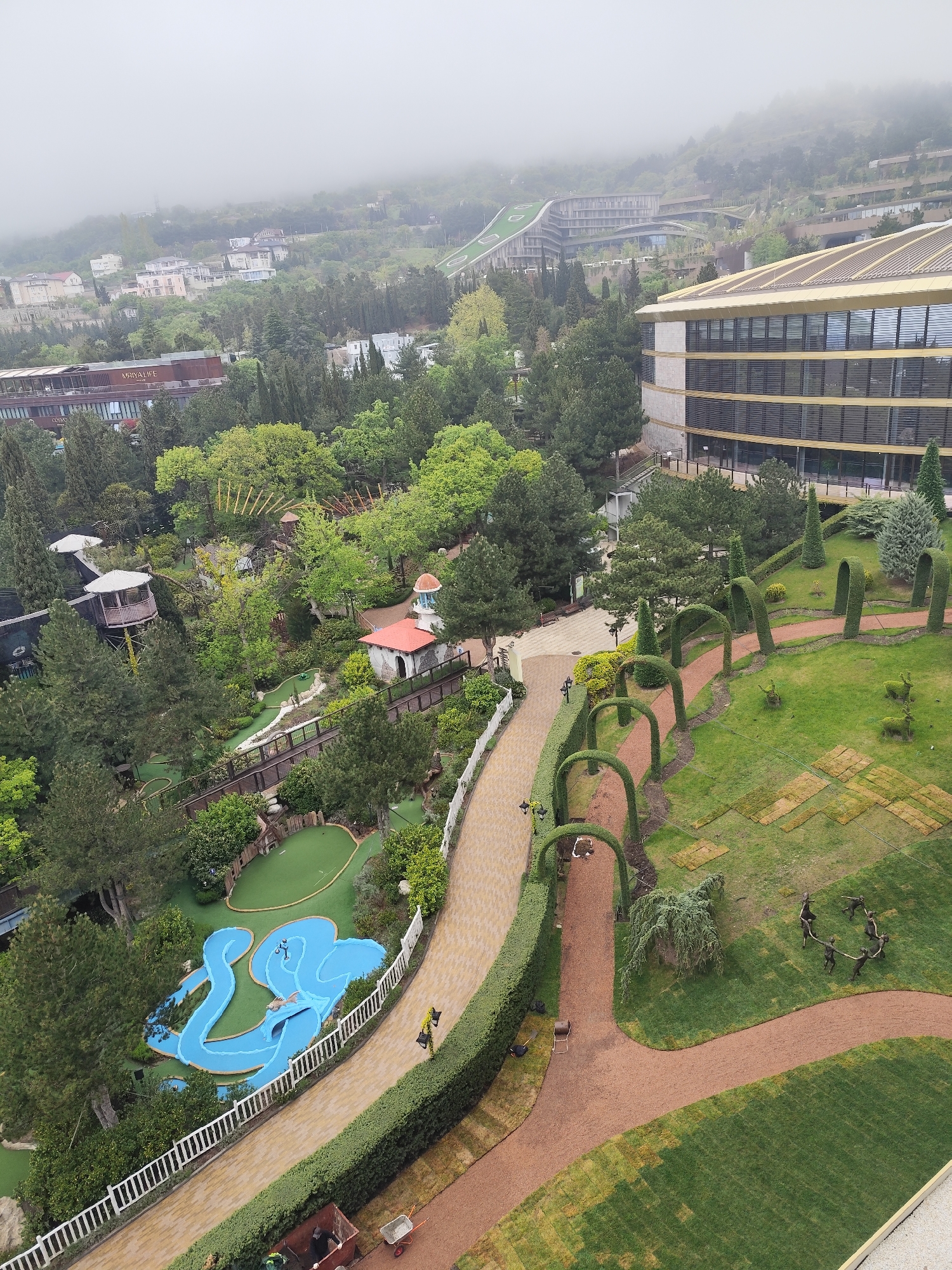 The image depicts an elevated view of a landscaped area in Симеизский поселковый совет, Ukraine. A paved pathway bordered by trimmed hedges and arched shrubbery cuts through a grassy lawn dotted with decorative elements like animal sculptures and figures in a ring. To the left, a miniature golf course features blue waterways and artificial green putting surfaces. Further back, a complex of buildings is visible, including a modern structure with a gold-colored roof. Trees and dense vegetation fill the space between the landscaped area and the buildings. In the background, a foggy or misty atmosphere obscures the distant hills and buildings. People are visible working on the landscape.