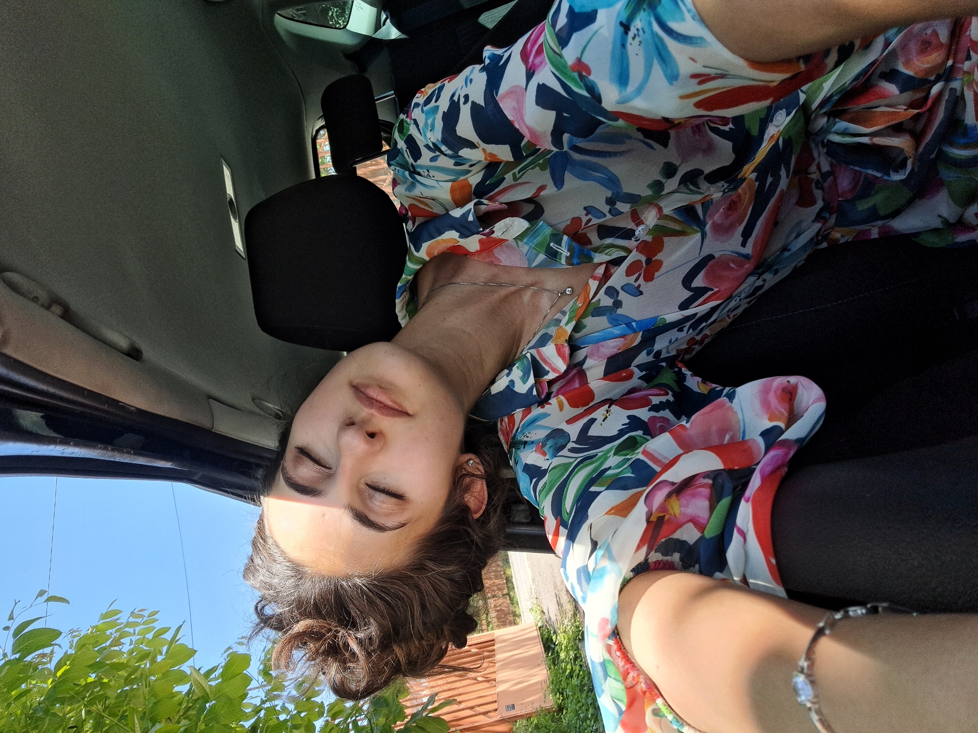 The image shows a woman with brown hair and closed eyes, wearing a floral blouse and a necklace, seated in a car. The interior of the car, including the headrest, is visible. Outside the car, there is a view of green trees and a blue sky. There is no information in the image to suggest this photo was taken in Sofia, Bulgaria.
