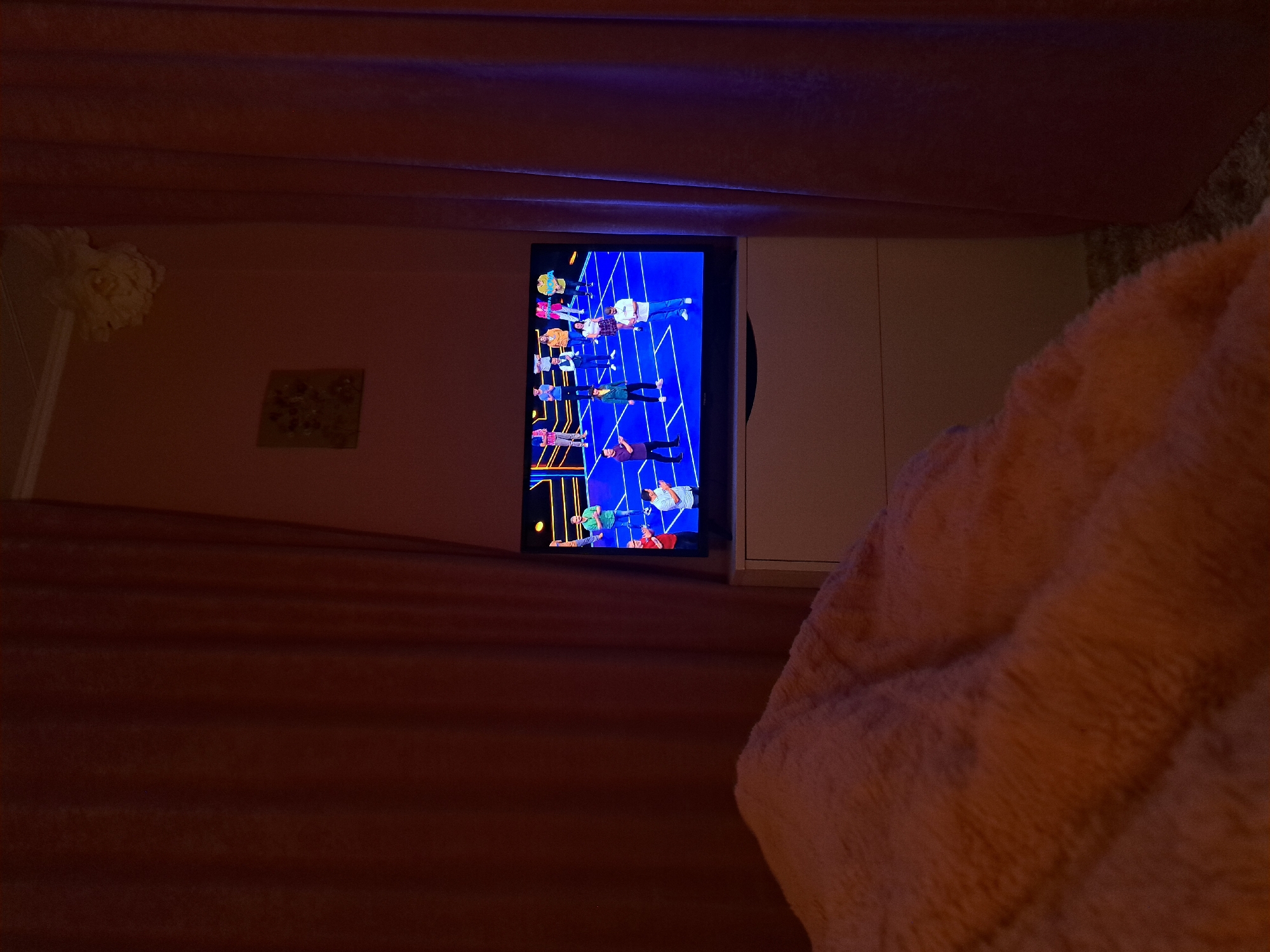 A television screen is displaying a colorful game show scene with multiple contestants standing on a set with blue and yellow accents. The screen is positioned above a white console or cabinet. The foreground is dominated by a soft, textured peach-colored blanket or cushion. The image appears to be taken in a dimly lit room, with a head figurine visible on the left near the edge of the wall. There is also a small framed picture on the wall. It is impossible to confirm the location of the city of Chepintsi, Bulgaria based on image data.