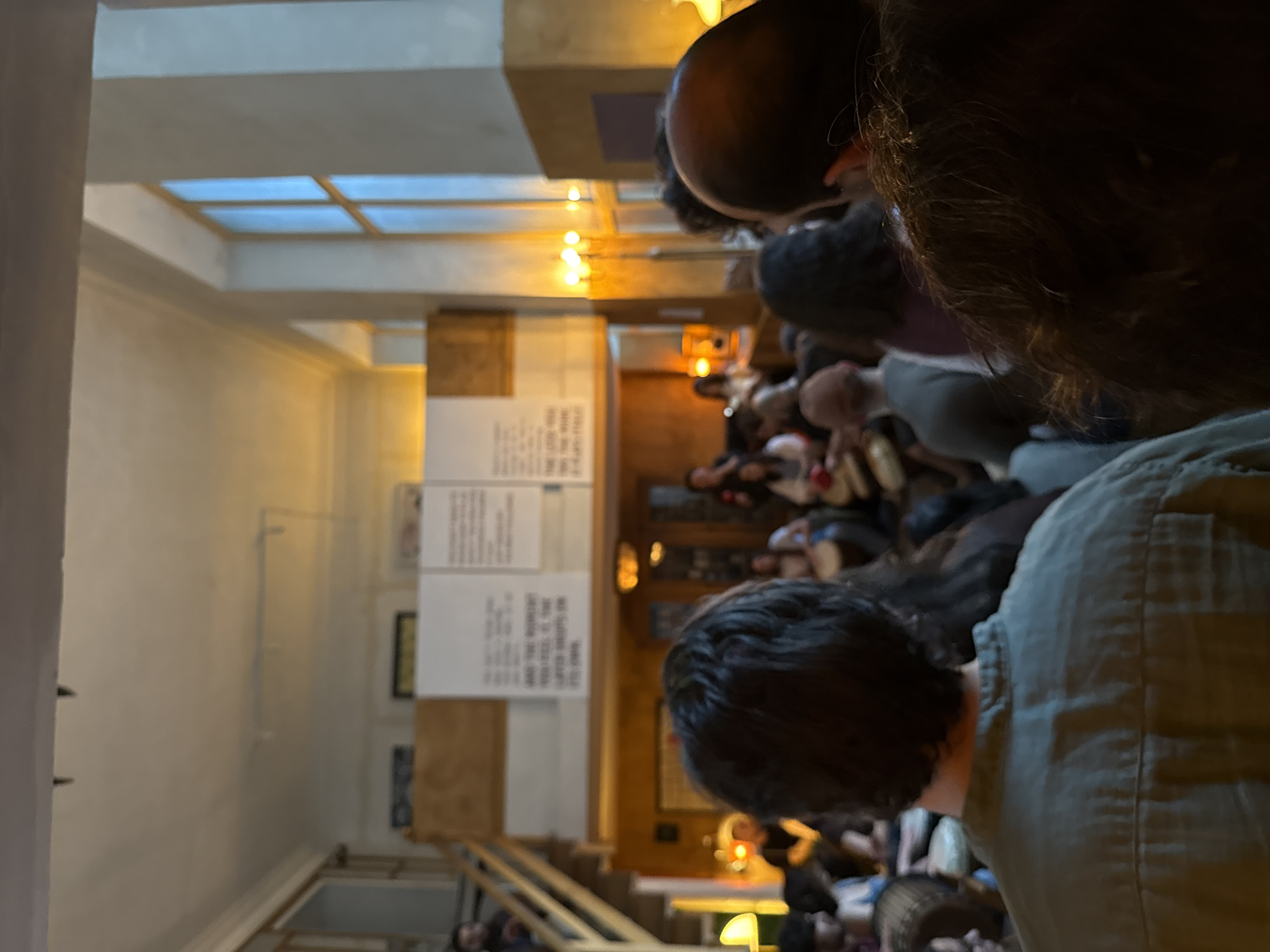 The image shows an interior view of a space, likely a lecture hall or auditorium, filled with people. The perspective is taken from the back of the room, looking towards a stage or presentation area. There are numerous individuals seated, with their heads and upper bodies visible. They seem to be attending an event or presentation.
In the foreground, several people are more clearly in focus. The person closest to the camera on the right wears an olive-green shirt. The lighting in the room is warm, with several overhead lights providing illumination.
On the left side of the frame, there are white boards with text written on them. Further back, there are decorative wooden accents and architectural details.
The city of Yerevan, Armenia is not evident from the given image and cannot be verified.