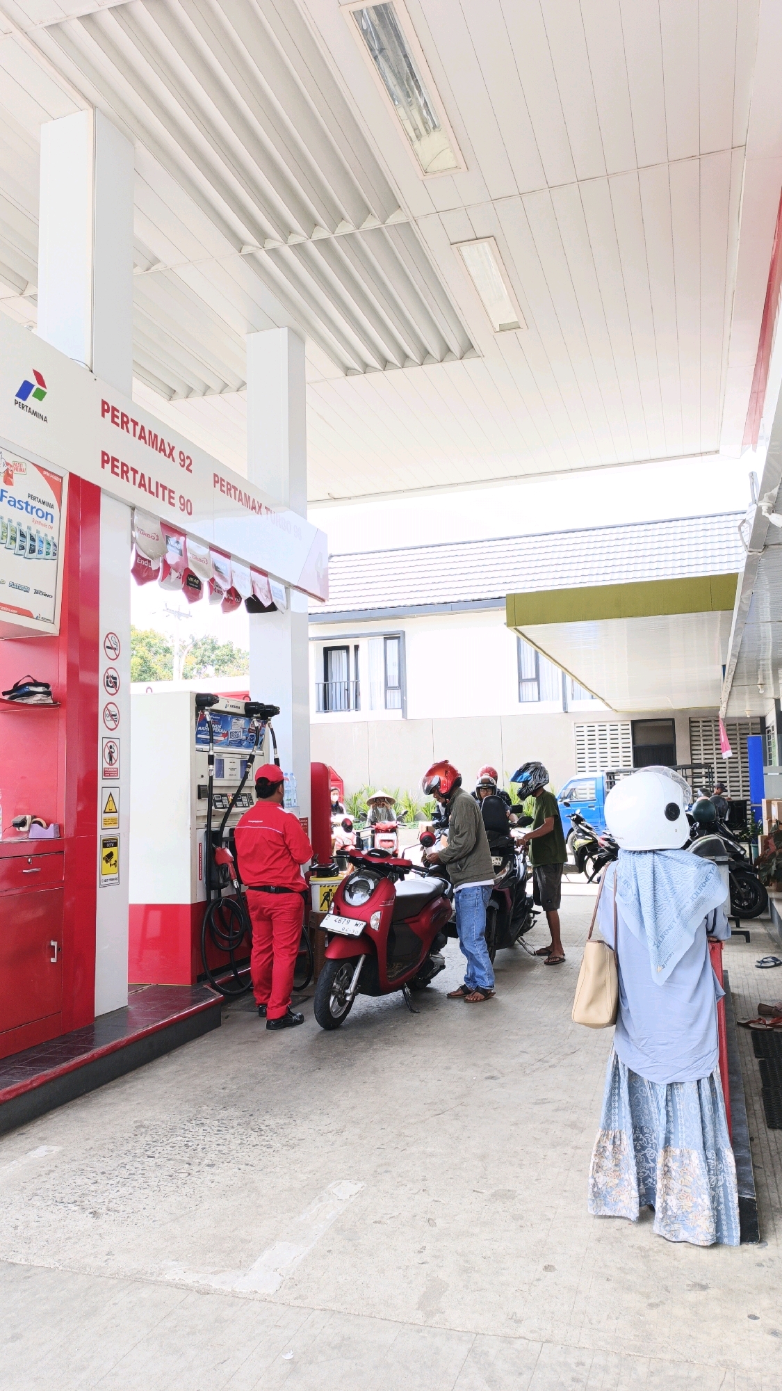 The image depicts a scene at a Pertamina gas station in Desa Wonoharjo, Indonesia. The gas station has a prominent red and white structure with signs indicating different fuel types, including "PERTAMAX 92" and "PERTALITE 90." Several people are present, mostly interacting with their motorcycles.
In the foreground, a person wearing a light blue hijab and a long blue and floral patterned skirt stands with their back to the camera, holding a beige bag. To their left, a red scooter is parked, and a man in a green jacket and blue jeans is attending to it. Behind him, another person in a dark helmet is also near a scooter.
To the left of the scooters, a gas pump is being operated by a man in a red uniform, presumably a gas station attendant. He is standing next to the pump, with his back to the viewer, and appears to be filling a scooter. More people and scooters are visible further back in the scene, some wearing helmets.
The environment includes the gas station's covered canopy with white ribbed panels and fluorescent lights. In the background, there is a building with a tiled roof and windows. The ground is paved with concrete. The overall atmosphere suggests a busy, everyday operation at the gas station.