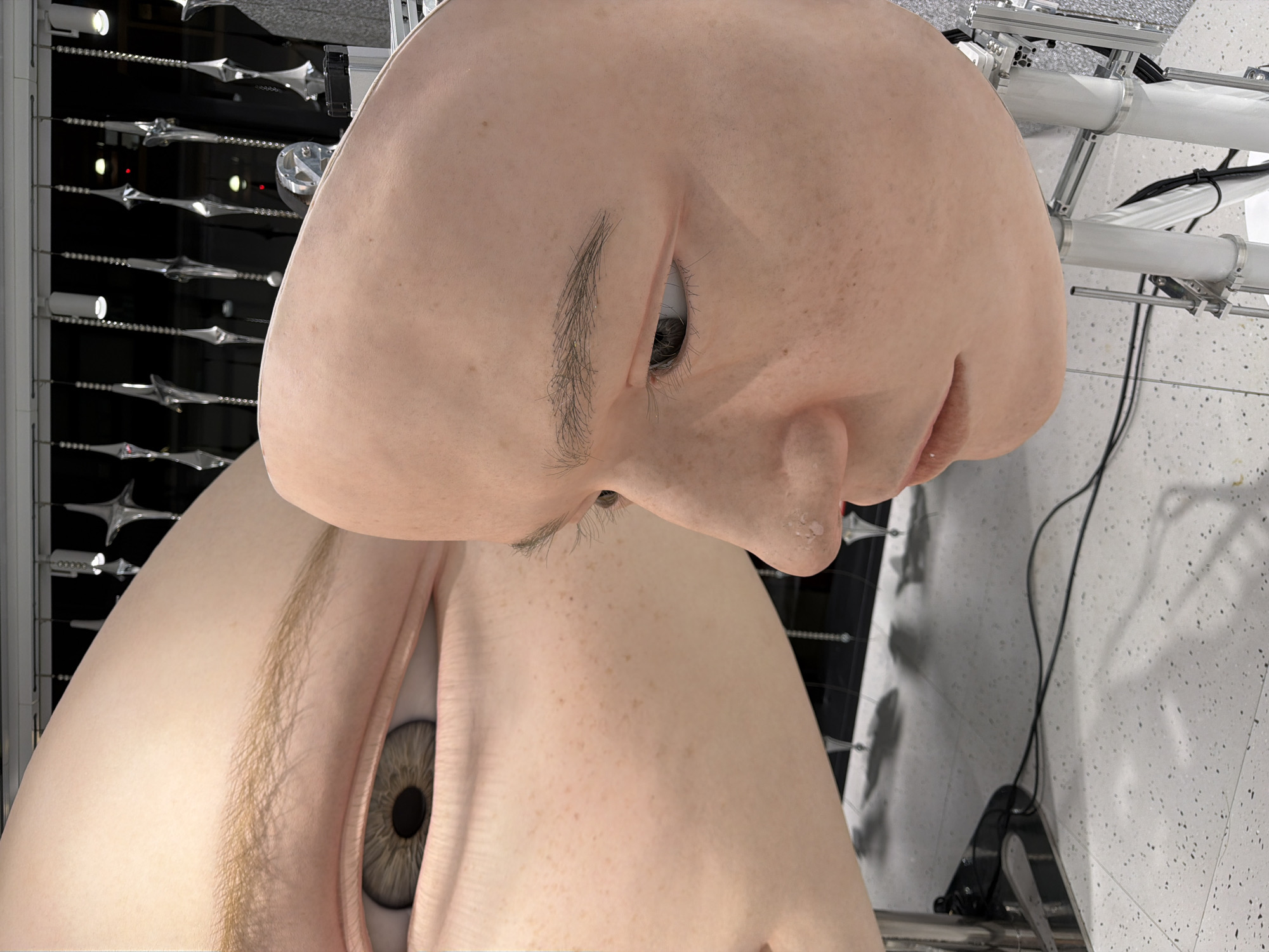 A close-up, angled view showcases two large, prosthetic faces, likely part of an art installation, set against a backdrop of dangling, metallic decorative objects. The face on the left is partially visible, focusing on its large, grey eye with a dark pupil and textured iris, and a portion of its forehead and cheek adorned with subtle freckles. The face on the right is positioned in profile, revealing its nose, lips, and a portion of its eye, also featuring freckles. The skin tones of both faces are light and peachy, with a smooth, matte finish. The background is dark, with the hanging decorations creating a sense of depth and reflecting light. The floor is a speckled grey and white tile. Metal poles and electrical wires are visible at the bottom of the frame, supporting the display. The overall impression is one of artificiality and a slightly unsettling realism, common in modern art exhibits. This installation is located at the GINZA SIX shopping complex in Chuo, Japan.