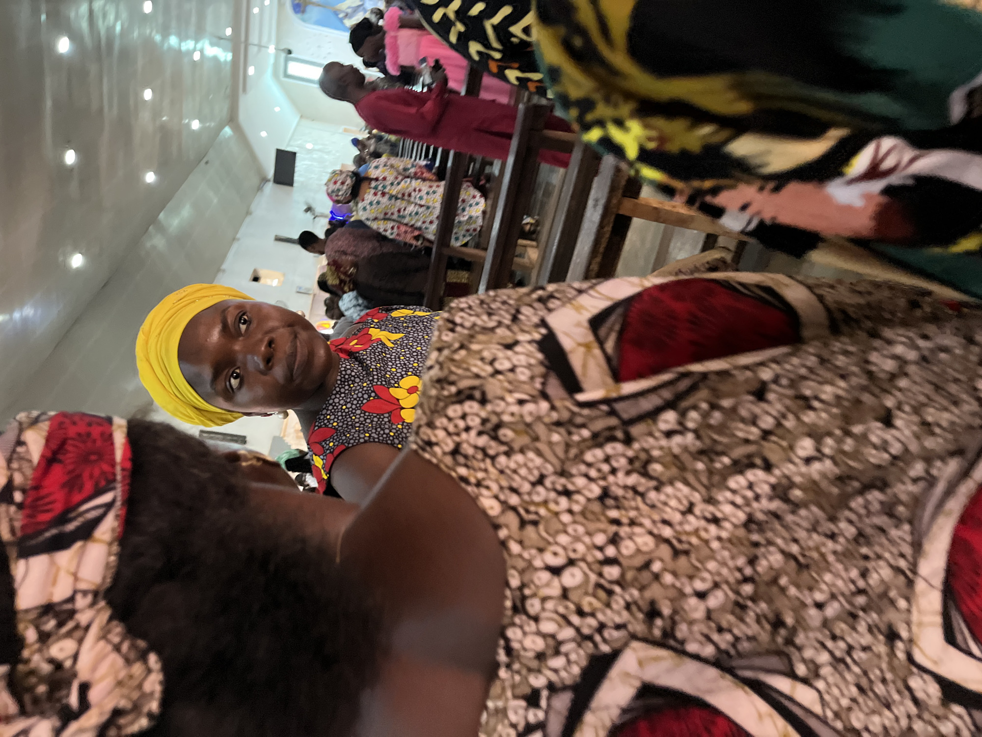 The image shows a group of people sitting in what appears to be a church in Jos, Nigeria. The camera angle is from a close-up perspective, focusing on a woman wearing a yellow headscarf and a patterned top. Other people are seated behind her, some dressed in colorful traditional African attire. The church interior has rows of wooden pews and bright overhead lighting. The image suggests a religious gathering or service.