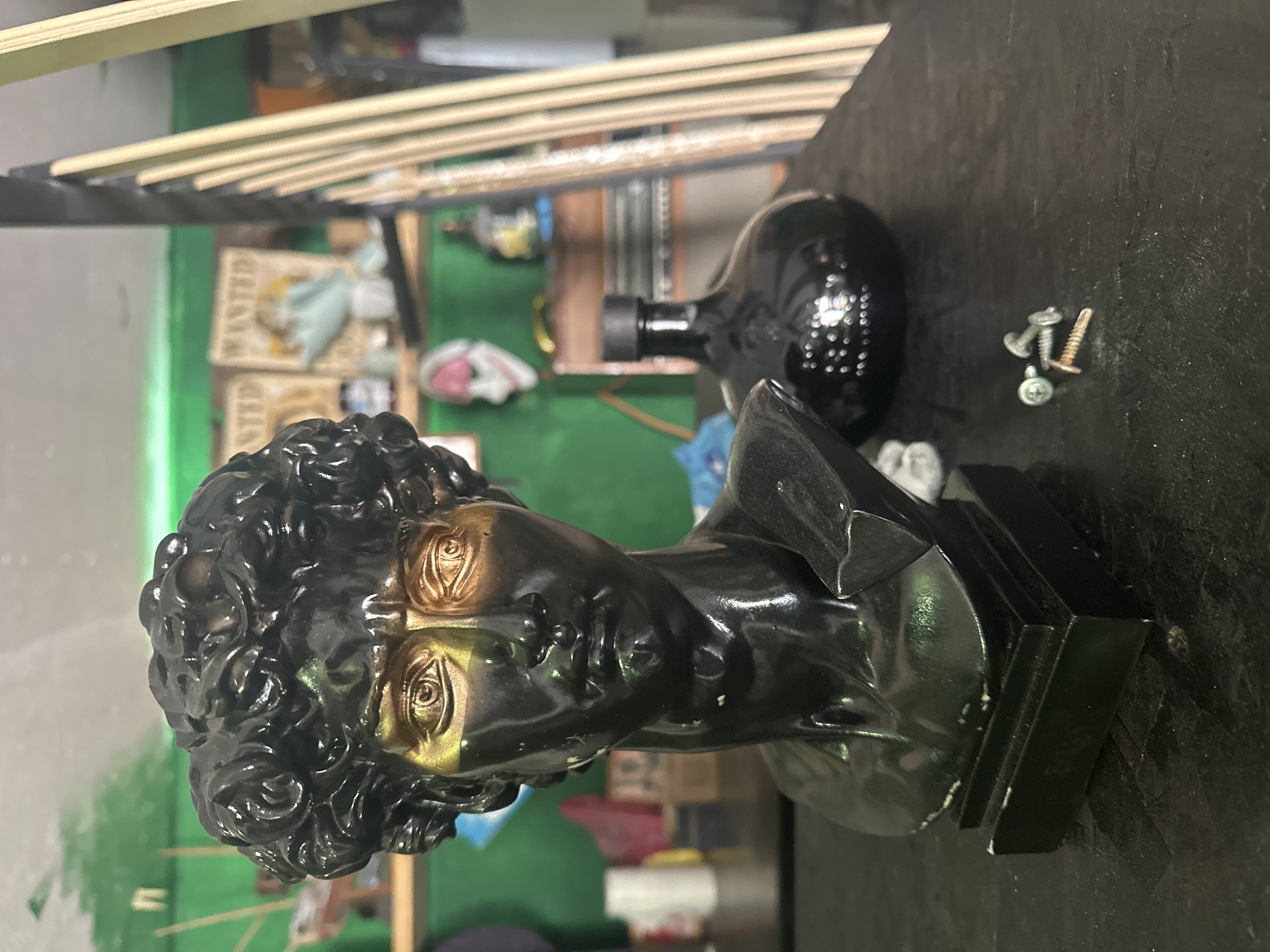 The image depicts a black and gold bust of a male figure, positioned on a dark, possibly black, surface. Next to the bust are a few screws. In the background, there's a bottle, wooden slats, and a green wall. The environment appears to be an indoor setting, possibly a workshop or storage area. The image doesn't provide enough information to determine whether it was taken in Andreyevka, Russia.