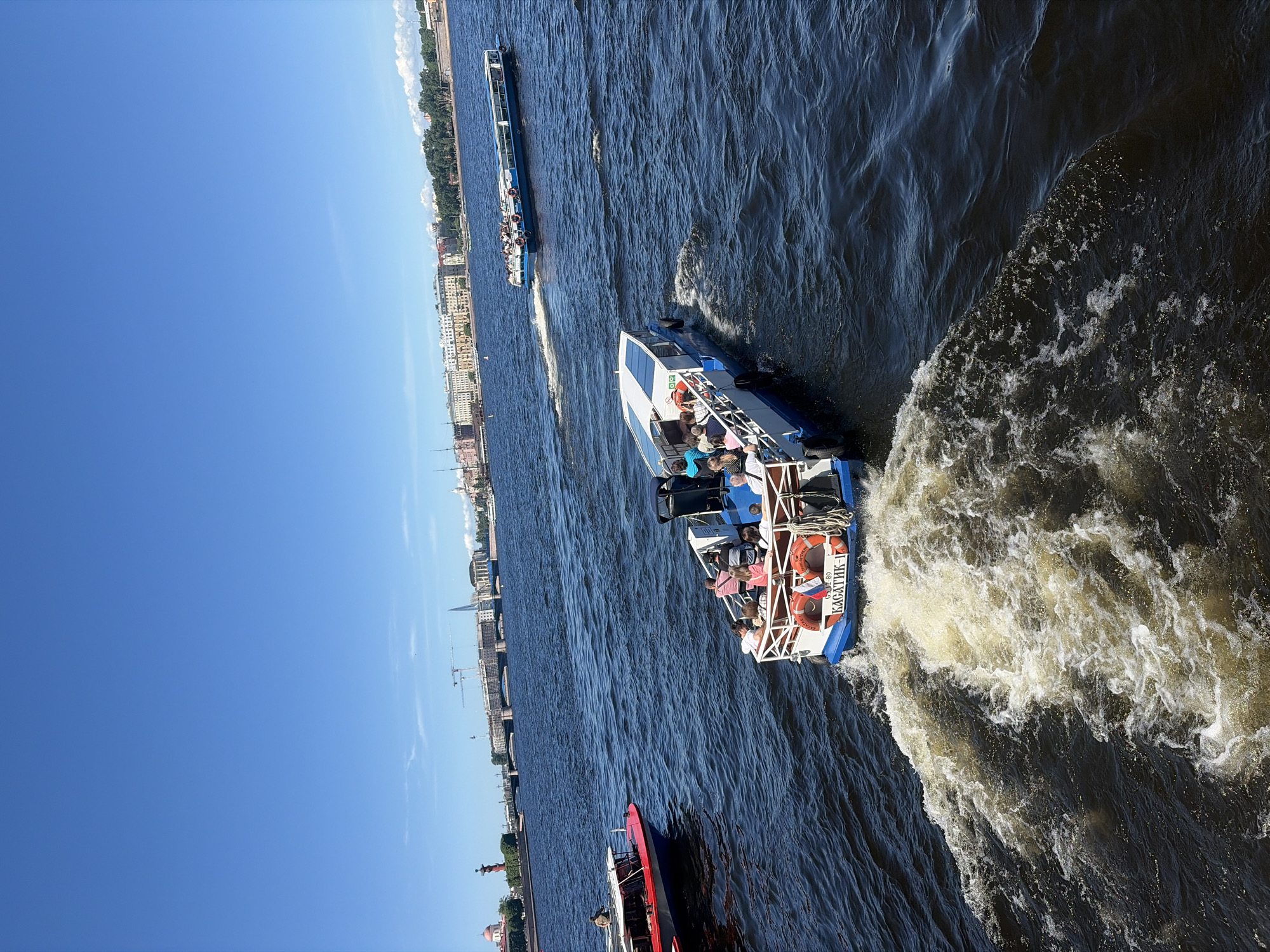 This image captures a vibrant, sunlit scene on the Neva River in St. Petersburg, Russia.
In the foreground, a white and blue passenger boat named "КАСАТИК-1" is moving away from the viewer, generating a prominent white wake. Several passengers, dressed in casual attire, are visible on its open upper deck, mostly seated. Two orange lifebuoys are secured to the stern railing.
In the midground, to the left, a section of another passenger boat, colored red and white, is partially visible. Further back and to the right, a longer blue and white passenger boat, also filled with people, travels in a similar direction, leaving its own wake. The water is a deep blue with ripples and foam from the boats' propulsion.
The background reveals a distinct St. Petersburg skyline. To the far left, a red Rostral Column and a red-domed building are clearly visible, marking the Vasilyevsky Island Spit. A large bridge spans the river in the middle distance. Beyond the bridge, various buildings line the horizon, including construction cranes and the tall, slender spire of the Lakhta Center skyscraper. To the right, a continuous stretch of classical European architecture along a tree-lined embankment defines the cityscape. The sky above is a clear, bright blue with a few faint cirrus clouds.