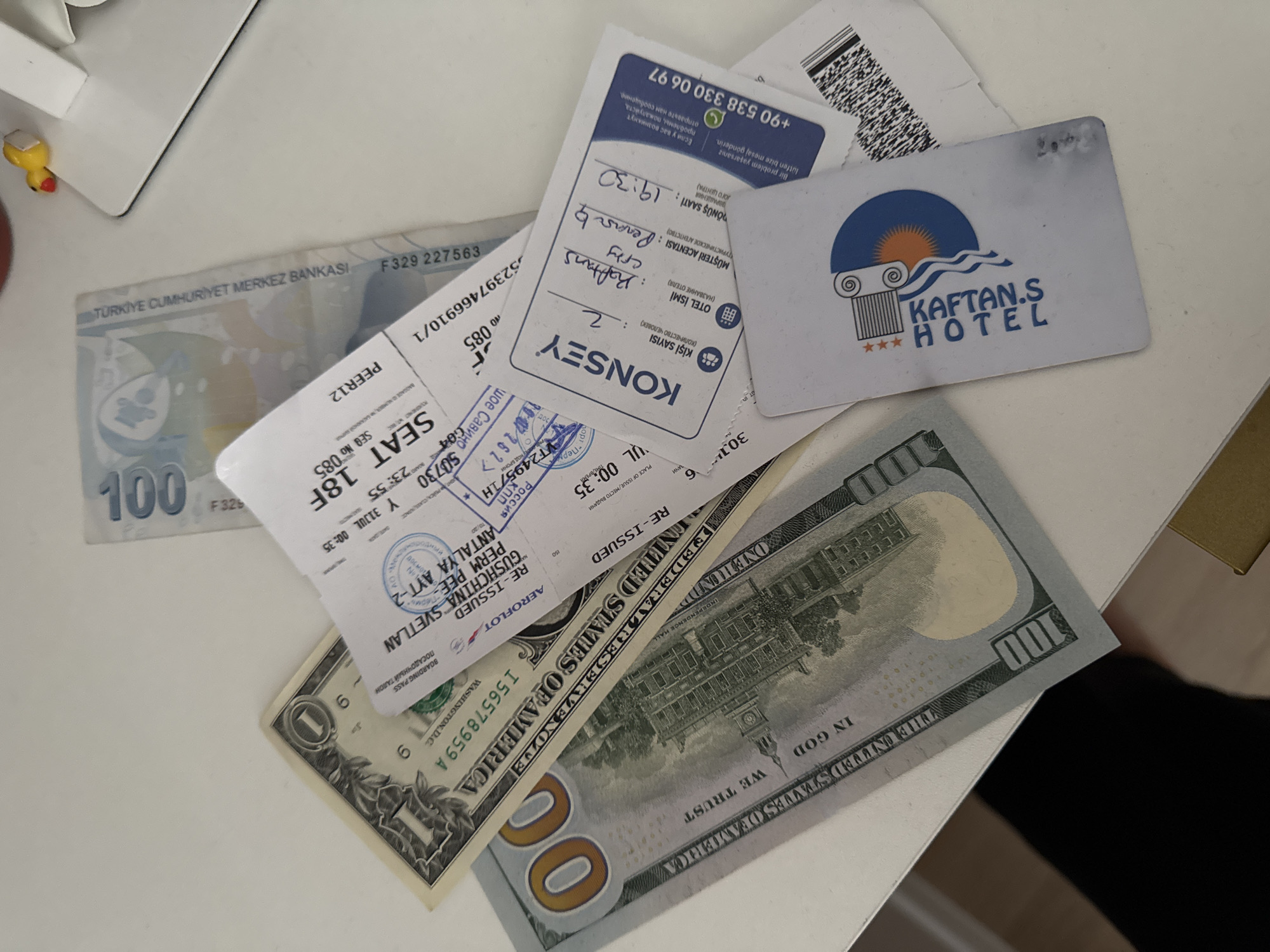 The image shows a table with several items, including US dollar bills, a Turkish Lira bill, a boarding pass, and a hotel key card. The boarding pass is for a flight from Antalya to Sweden. The hotel key card is for Kaftans Hotel. The scene appears to be indoors, possibly on a desk or table, given the flat surface and white background. There is no indication of the city of Perm, Russia in the image.