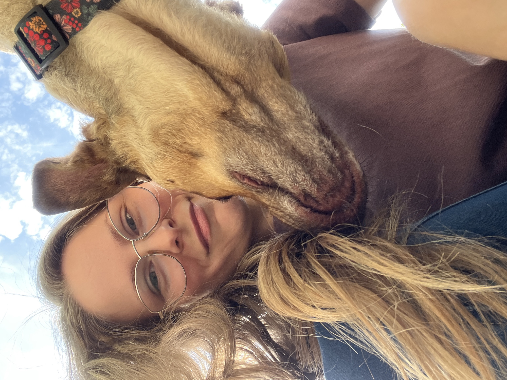 In a close-up selfie, a fair-skinned woman with blonde hair, wearing round wire-rimmed glasses, smiles at the camera. Her hair is partially obscuring her face. A light brown dog is nuzzling her cheek, with its fur visible against her skin. The dog is wearing a black collar with a colorful floral pattern. The background shows a bright sky with scattered white clouds. The woman appears to be wearing a maroon t-shirt. The image captures a moment of affection between a woman and her dog.