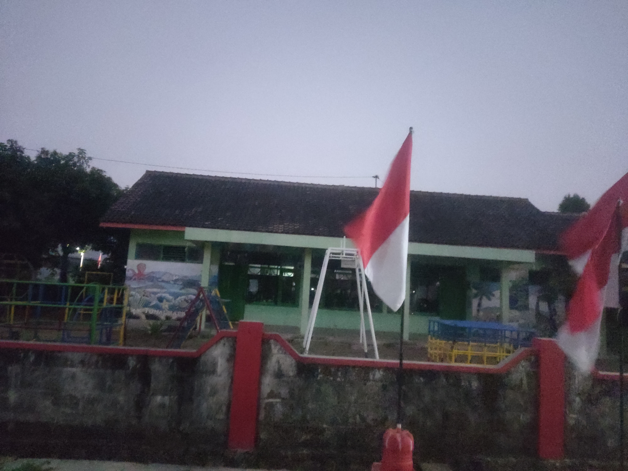 The image captures a scene at dusk in Trihanggo, Indonesia, featuring a school building and its outdoor area. A long, single-story building with a dark-tiled roof and green walls dominates the background. The building appears to be a school, as suggested by the playground equipment visible in front of it. This equipment includes colorful metal structures with climbing frames and slides, likely for children's recreation. Two Indonesian flags, red and white, are prominently displayed, one on the left and another on the right, with one of them waving in the breeze. The flags are mounted on poles and are positioned in front of the building and a low, weathered stone wall. The wall has red vertical pillars and a red top railing. The sky is overcast, indicating either early morning or late evening. Trees are visible to the left of the building. The overall impression is of a quiet, communal space, possibly a primary school, during a time of low light.