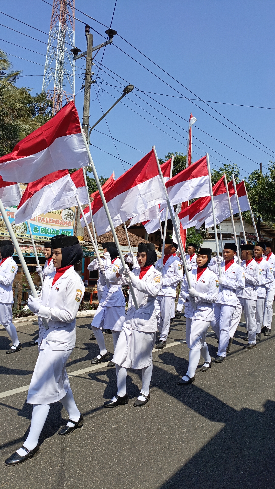 A group of young people in white uniforms and black head coverings march in formation on a paved road, holding Indonesian flags. The flags are red and white, with the red stripe on top and the white stripe on the bottom. The marchers are wearing white jackets with red scarves and white skirts or pants. They are also wearing white socks and black shoes. The march is taking place in a sunny outdoor environment, with buildings and trees visible in the background. A tall radio tower is also visible in the distance. The location is identified as Krajan Lor, Indonesia.