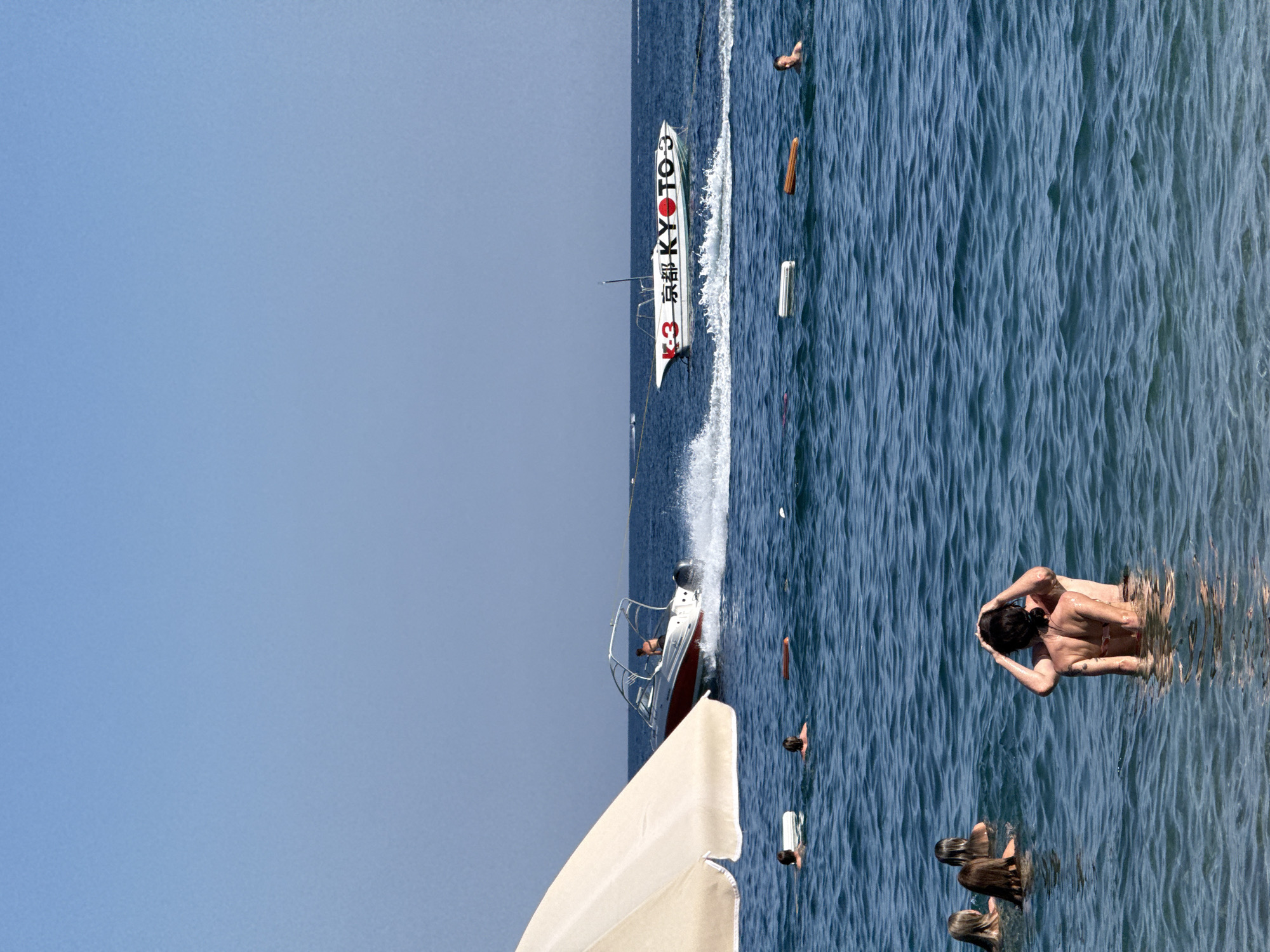 This is a photograph of a lively scene at sea, likely during daytime given the bright sunlight. The image captures people enjoying a day on the water.
The primary subjects are several individuals swimming and interacting in the ocean. In the foreground, a couple embraces in the water, appearing relaxed and content. Further out, other swimmers are visible, some with their heads above water, suggesting recreational swimming or wading.
A prominent object in the mid-ground is a white speedboat with red accents, moving at a moderate speed and leaving a wake. The boat has "KYOTO" and what appears to be Japanese characters printed on its side, along with the number "3". There are also what look like buoys or markers in the water. In the lower left of the image, a portion of a large, light-colored beach umbrella or canopy is visible, hinting at a beach or boat-based leisure setting.
The weather appears to be sunny and fair, with clear blue skies and calm, blue waters. The rippling surface of the water indicates some gentle movement. The overall mood is one of summer recreation and enjoyment.
The visible text on the boat reads "KYOTO 3" and includes Japanese characters that appear to be "京都" (Kyōto).
The location context of Budva, Montenegro, suggests a coastal resort area known for its beaches and opportunities for water activities.