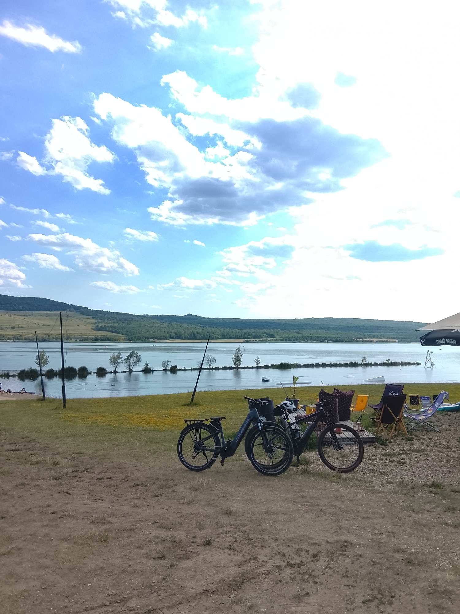 The image depicts a picturesque lakeside scene on a sunny day. The foreground features two electric bicycles parked on a sandy patch of ground. Behind the bicycles, a grassy area is dotted with several colorful lounge chairs and some scattered items, suggesting a recreational area.
The middle ground is dominated by a calm, expansive lake. The water reflects the bright blue sky, which is adorned with fluffy white clouds, indicating fair weather. Along the lake's edge, a line of trees and reeds creates a natural barrier between the water and the grassy shore. In the distance, a gently sloping hillside covered in lush green forest forms the backdrop. Several wakeboarding ramps are visible in the water, hinting at the type of activities that might take place here.
A few indistinct figures can be seen on the far side of the lake, near the water's edge, suggesting the presence of other visitors. The overall atmosphere is serene and inviting, perfect for outdoor leisure. The time of day appears to be midday or early afternoon, given the bright sunlight and the shadows. A partial view of an umbrella on the right side of the frame reveals the text "OBRAT PROUD" printed on it.