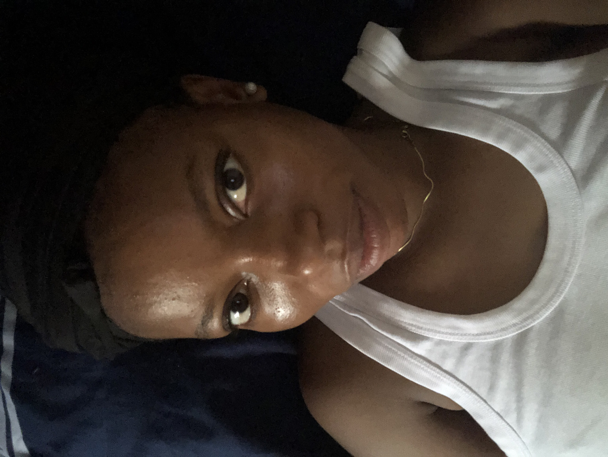 A close-up, angled shot of a person with dark skin and brown eyes, wearing a white tank top and a thin gold chain necklace. The person is lying down on what appears to be dark blue bedding with some light blue/grey stripes visible. The lighting is dim, with the right side of the person's face more illuminated. A single earring is visible on the upper ear of the subject. The shot is tightly cropped around the head and upper chest area of the person. There is no contextual information related to the city of Kashere, Nigeria, or any other location within the image.
