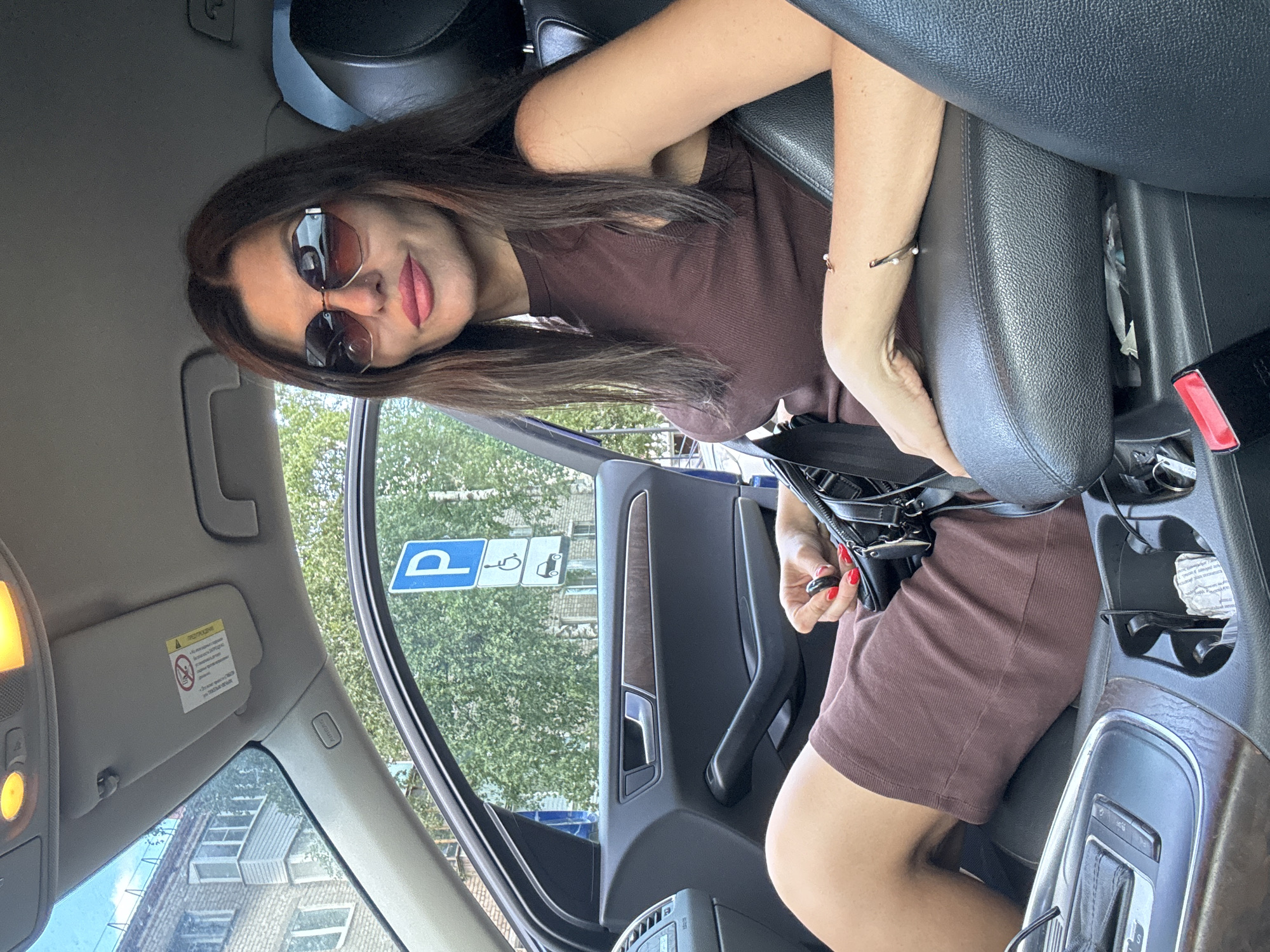 A woman is sitting in the driver's seat of a car, wearing sunglasses and a brown sleeveless dress. She has dark brown hair and is wearing red nail polish. She is holding a black handbag and a car key. The car appears to be parked on a street in Chaikovsky, Russia, as indicated by the presence of a parking sign with Russian text. The background shows a brick building and trees.