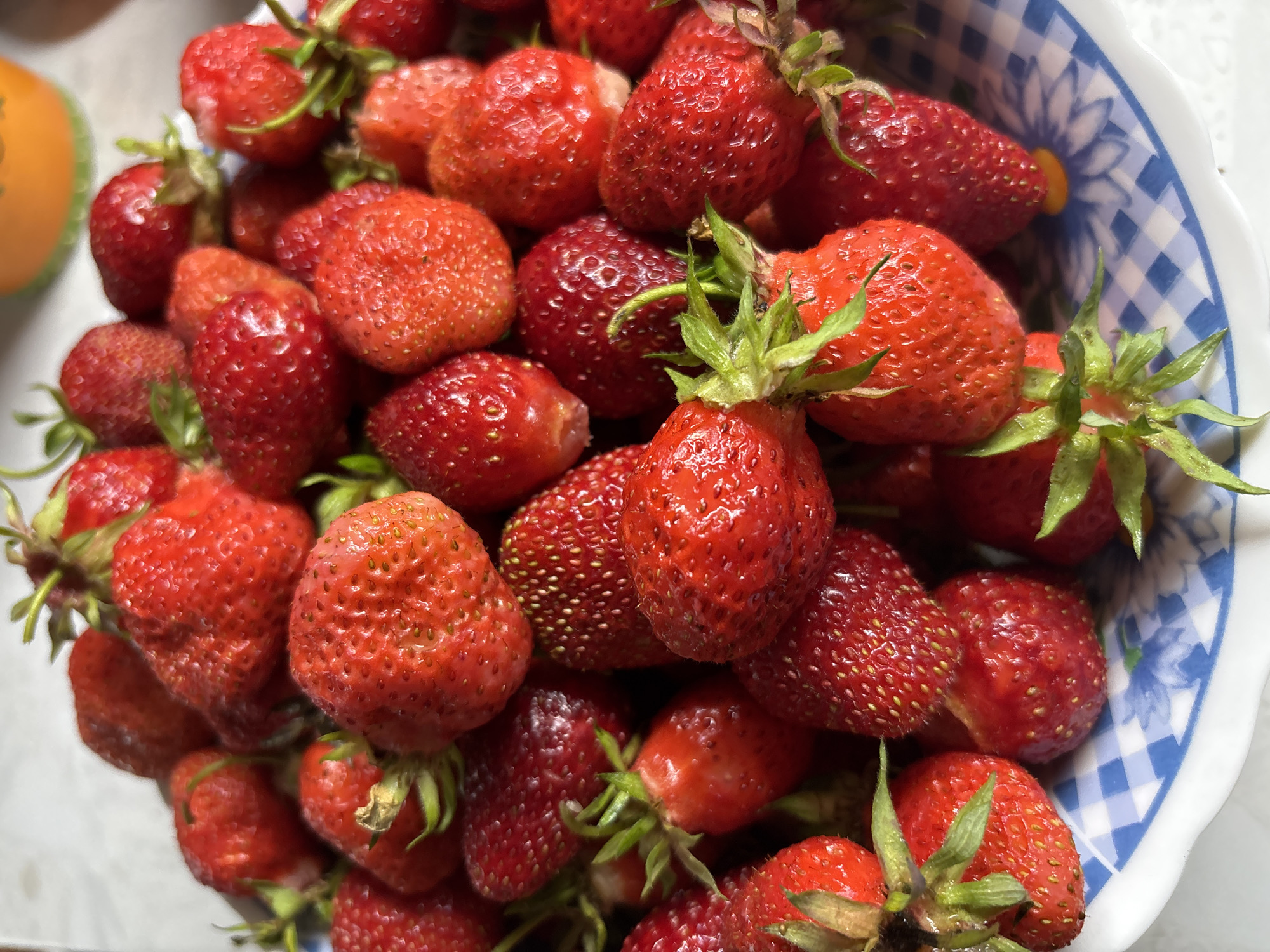 The image shows a close-up, overhead view of a white bowl with a blue and white checkered pattern filled with fresh strawberries. The strawberries are a vibrant red, with visible seeds and green stems. The bowl appears to be on a light-colored, textured surface, possibly a tablecloth or countertop. In the upper right background, a portion of an orange container with a green rim is visible, out of focus. The scene is well-lit, suggesting natural light. There are no people or animals present. The image does not provide any information that would indicate its location in the city of Кордово, Russia.