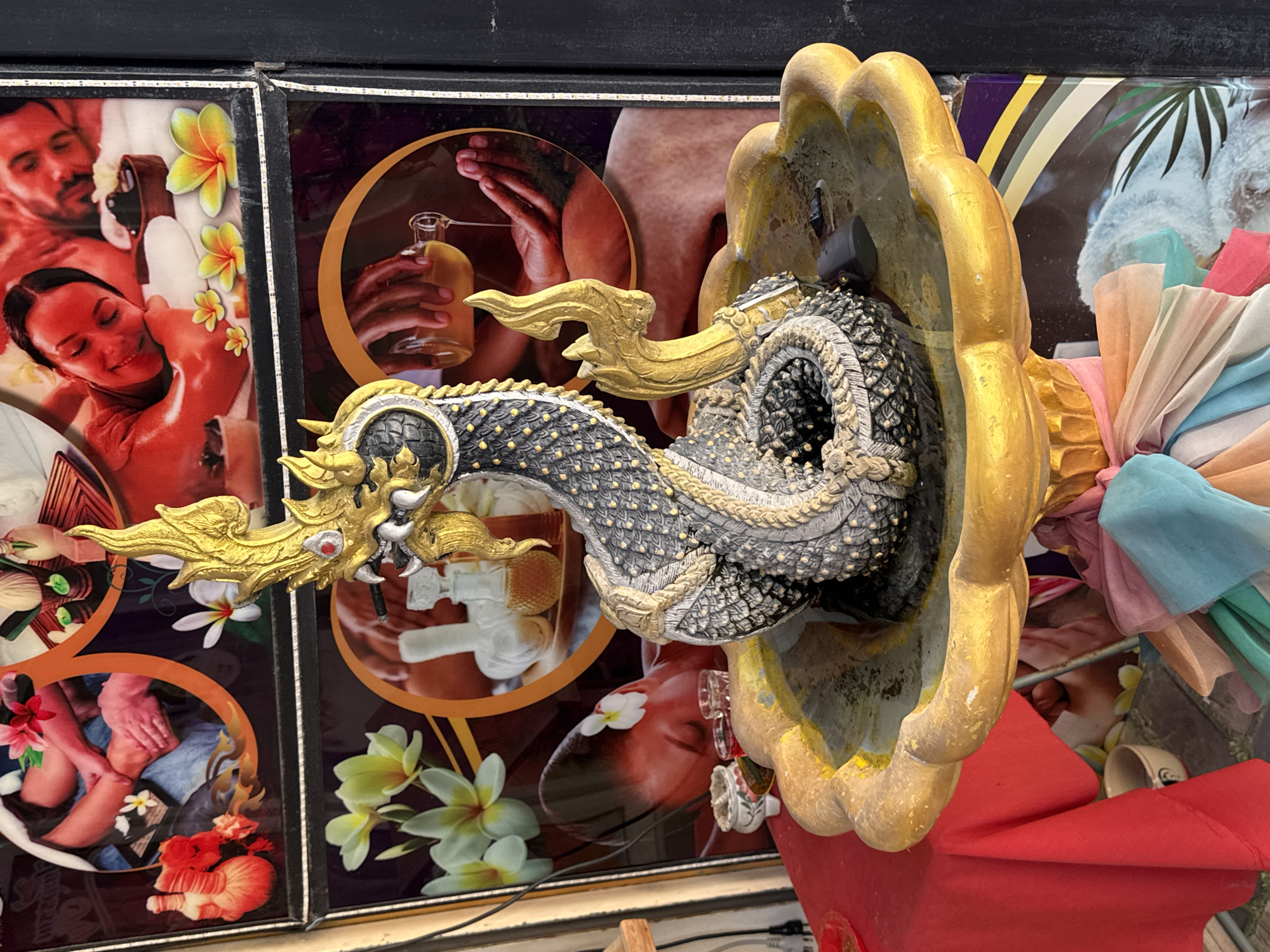 In Patong, Thailand, a fountain featuring a serpentine statue with ornate golden accents is placed in front of large posters depicting spa and massage scenes. The statue, detailed with scales and a dragon-like head, is centrally positioned within a decorative golden basin filled with murky water. A small black device is visible submerged in the water. The background posters showcase various aspects of relaxation and wellness, including close-ups of hands receiving massage, aromatherapy elements like oils and herbs, and couples enjoying spa treatments. The overall scene is vibrant, with colorful fabrics draped around the base of the fountain, suggesting a commercial establishment, possibly a spa or a shop catering to tourists. The lighting is bright, indicating daytime.