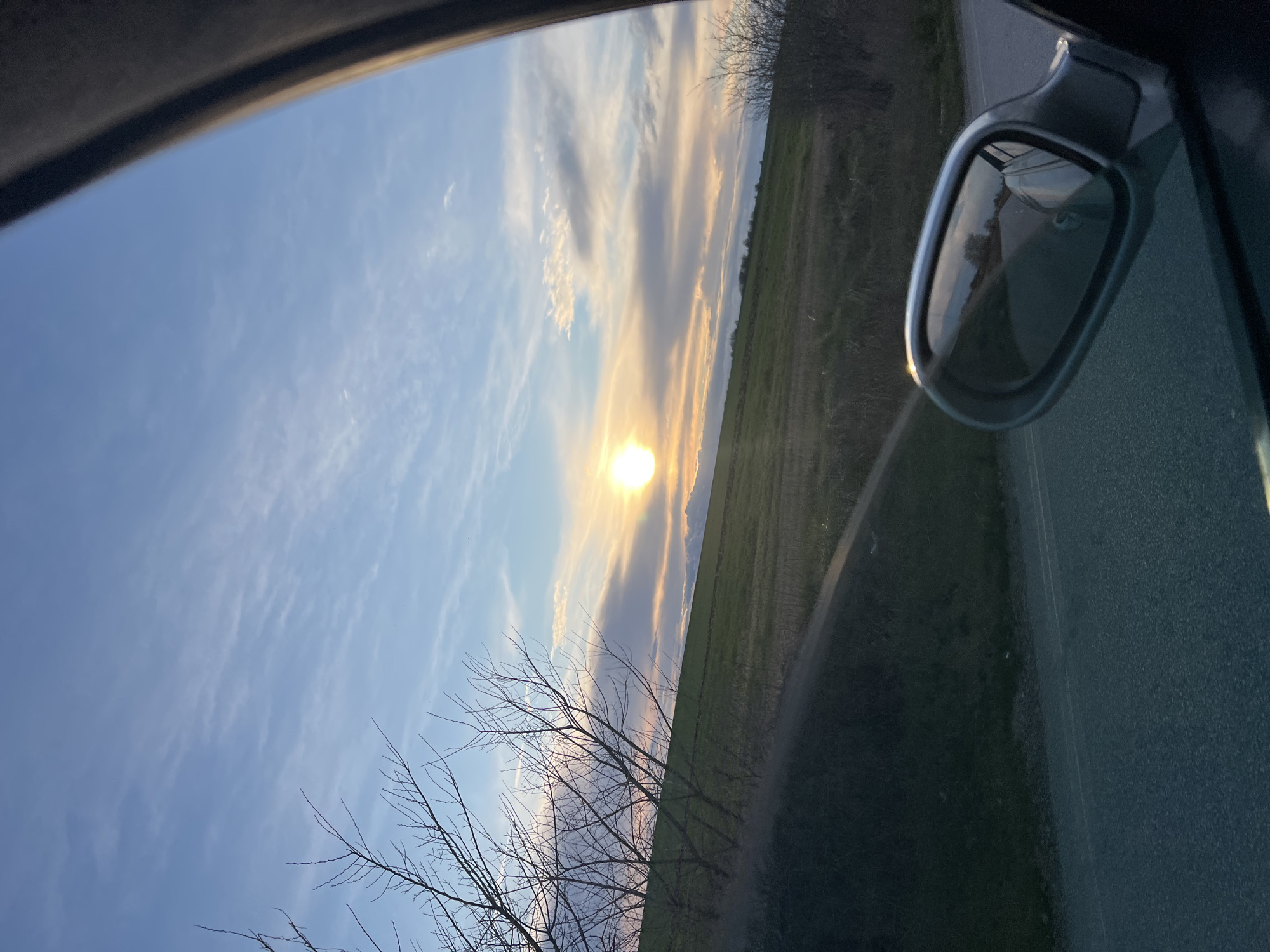 The image depicts a landscape as seen from inside a vehicle, possibly a car. The sun is setting or rising, casting a bright glow near the horizon. The sky is a mix of blue and hues of orange and yellow, with thin, streaky clouds. The landscape appears to be a field, possibly agricultural land, with hints of green. Bare trees or shrubs are visible near the bottom of the frame. Part of the car's side mirror is visible in the upper right, reflecting the scene. The rest of the interior and exterior car elements frame the shot.
Given the specified location of Zhitnitsa, Bulgaria, it's possible the landscape is typical of the region. However, without further context, no specific landmarks or features can be identified to confirm the exact location. No people are visible in the image. The primary action is the passive observation of the landscape, and the main subject is the natural environment at sunrise/sunset.