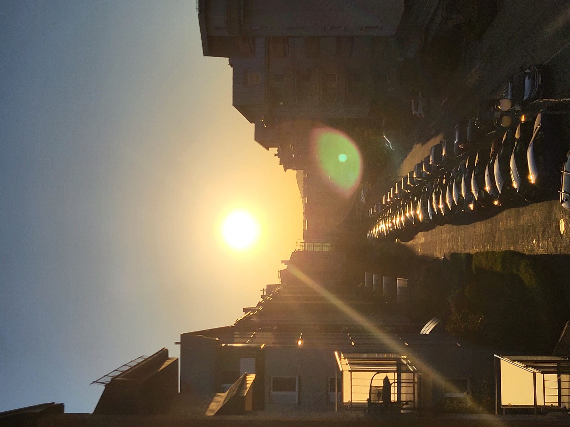 Here's a detailed description of the image:
**Setting:** The image depicts an urban landscape at either sunrise or sunset. The scene is dominated by architectural structures and rows of parked vehicles.
**Objects:**
* **Buildings:** The architecture includes both high-rise apartment buildings and what appear to be lower residential or commercial buildings.
* **Vehicles:** A long row of parked cars is prominently visible, oriented in a line along the side of the buildings. These are likely residential cars.
* **Sun:** A bright sun is centrally positioned, casting a strong light and creating lens flare.
**Environment:**
* The sky shows a gradient of colors, ranging from light blue to orange, which would be expected during sunrise or sunset.
* Vegetation is apparent along the edges of buildings, indicating a green space or garden.
**Composition:**
* The image is oriented vertically.
* The overall tone is warm due to the sunlight.
**Location:** Without further information or metadata, the exact location of the photo in the city of Unknown, Unknown, is impossible to pinpoint.