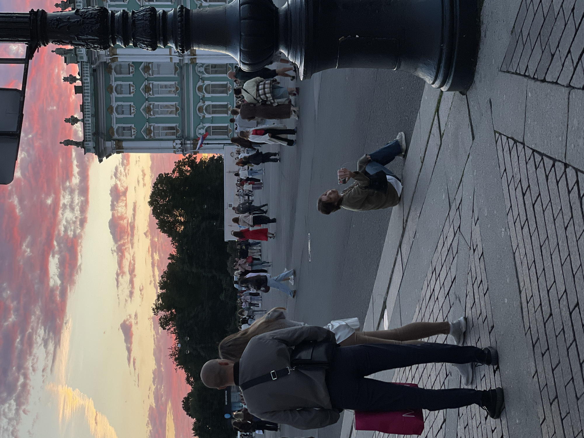 The image captures a street scene in front of a grand, light teal building with white trim and ornate architectural details, illuminated by a sunset with vibrant pink and orange clouds. In the foreground, a man stands with his back to the camera, wearing a gray jacket, dark pants, and a crossbody bag, holding a bright pink shopping bag. To his left, a woman in beige pants and a white top stands partially obscured. Further into the scene, a woman is seated on the curb, looking at her phone. A large, dark lamppost is prominent on the right side of the frame. In the background, numerous people are walking and congregating on a paved area, with trees and the building forming the backdrop.