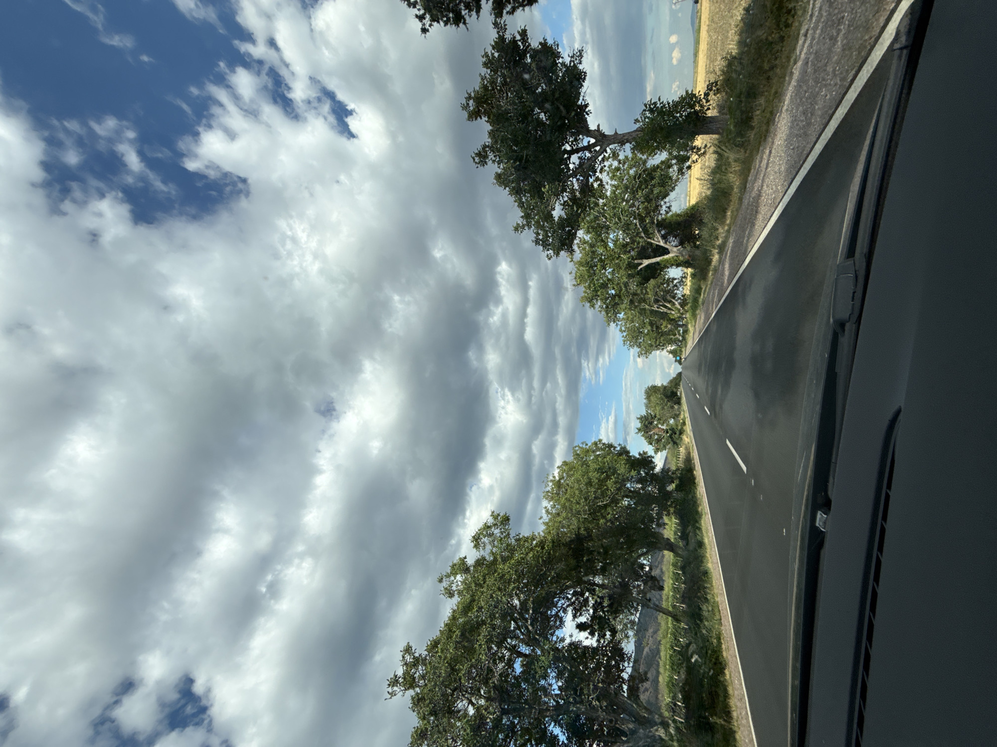 The image depicts a rural road winding through a landscape with a dramatic sky. The road is asphalt, with a white line marking the lane. On either side of the road are mature trees with lush green foliage, casting shadows. Beyond the trees, the land appears to be a mix of cultivated fields and natural vegetation, with rolling hills in the distance. The sky is a prominent feature, with a mix of heavy, dark clouds and patches of bright blue sky, suggesting dynamic weather conditions. The perspective is from inside a vehicle, looking out over the dashboard and windshield. The scene is set in Kaloyanovo, Bulgaria, a town known for its agricultural landscape. There are no people or animals visible in the image. The overall impression is one of a journey through a scenic, rural environment under a changeable sky.