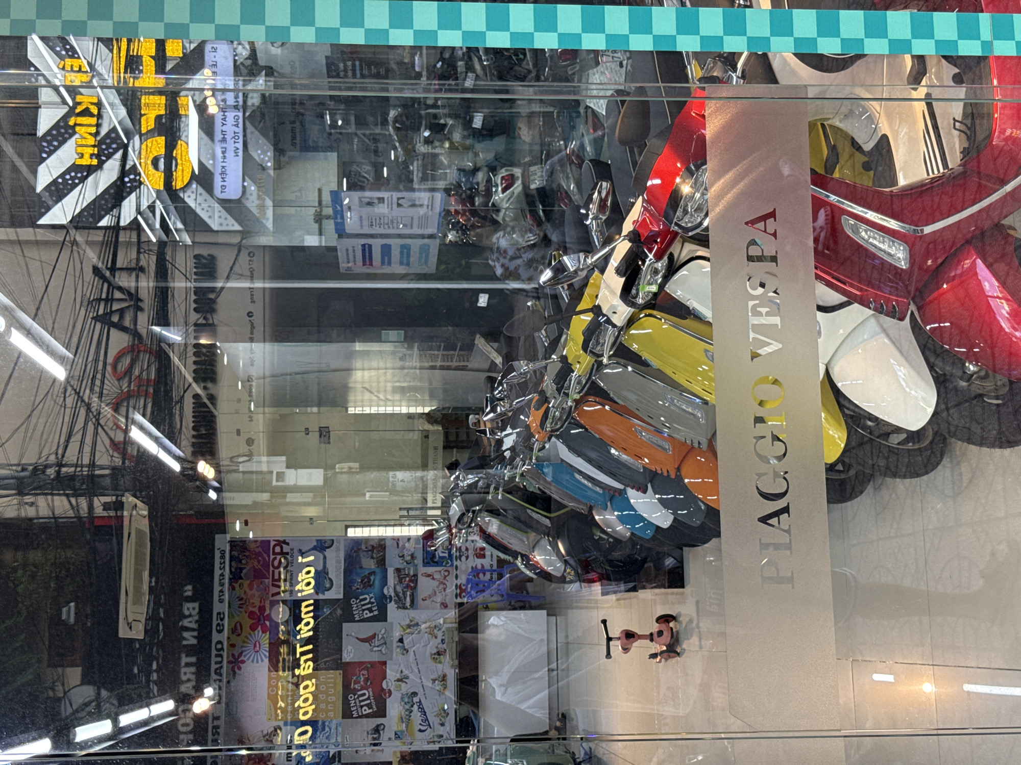 The image captures a street-level view of a Piaggio Vespa dealership, likely located in Nha Trang, Vietnam, as suggested by the Vietnamese text on signs. The scene is dominated by reflections on a large glass storefront, creating a layered and somewhat abstract composition. Inside the dealership, several Vespa scooters are parked in rows, displaying a variety of colors including red, yellow, white, blue, orange, and gray. A pink child's scooter is visible on the left side of the frame, near the entrance.
The reflections on the glass show overhead power lines and wires, signs with Vietnamese text, and what appear to be promotional posters featuring the Vespa brand. One prominent sign inside the store, partially obscured by reflections, reads "PIAGGIO VESPA". On the exterior of the glass, a large frosted decal also displays "PIAGGIO VESPA". The lighting suggests it is daytime, with artificial lights visible inside the showroom and natural light contributing to the reflections. The overall impression is one of commercial activity and the display of merchandise through a transparent barrier.