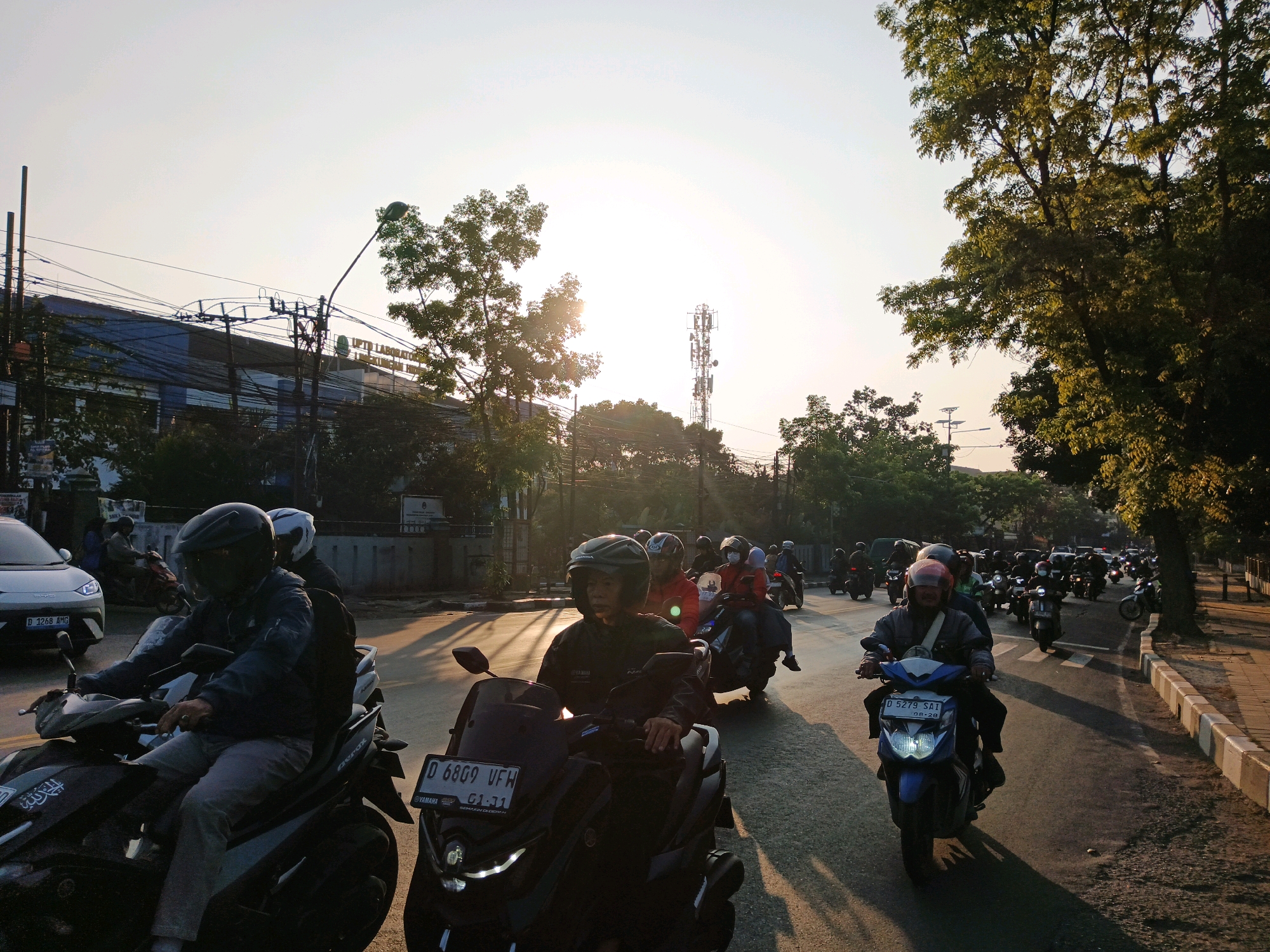 The image depicts a street scene in Bandung City, Indonesia, during what appears to be late afternoon or early evening, judging by the warm, low-angle sunlight casting long shadows. The foreground is dominated by a procession of motorcycles, with riders wearing helmets. One motorcycle in the immediate foreground has the license plate "D 6809 UFW". Several other motorcycles are visible behind it, some with multiple passengers.
The street is paved and shows tire marks. To the left of the street, there is a sidewalk and a road with a white car on it. Behind the road, buildings and trees are visible. One building has signage that is partially obscured but appears to read "UPTD LABORATORIUM..."
Further into the background, a cell tower stands tall against the sky, and more trees line the right side of the road. The sky is clear with a bright, hazy quality near the horizon, indicating the sun is low. The overall impression is of moderate urban traffic on a day with clear weather.