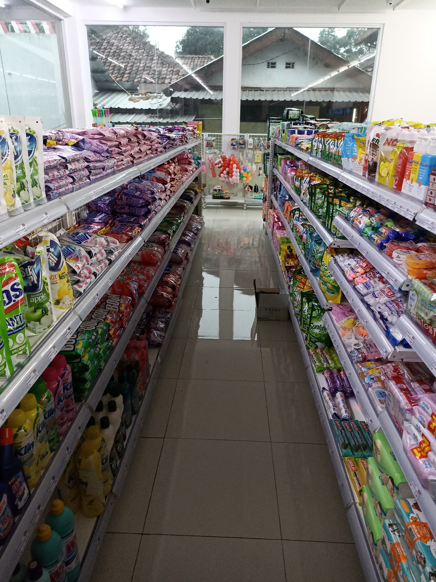 This image depicts the interior of a small convenience store or minimarket located in Tompokersari, Indonesia. The perspective is from within the store, looking down an aisle towards the front windows, which offer a view of outdoor roofing and trees.
The aisle is lined with shelves on both sides, fully stocked with various household and personal care products. The left side of the aisle displays numerous bottles and pouches of cleaning supplies, detergents, and possibly dish soap. Notable brands or packaging colors include green, yellow, and blue. Further down, there are rows of what appear to be laundry detergent packets in shades of purple and pink.
The right side of the aisle also features shelves stocked with a variety of items. Prominently displayed are packages of what seem to be toiletries, possibly soaps or cleansing wipes, in colorful packaging. There are also rows of smaller, individually wrapped items, and some larger bags of products. Towards the back of this aisle, near the front windows, there are shelves with colorful toys or decorative items, and some packaged goods with bright labels.
The floor is tiled with light-colored square tiles. The lighting in the store appears to be a mix of overhead fluorescent lights and natural light filtering in from the windows. A discarded cardboard box is visible on the floor in the middle of the aisle. The overall impression is of a well-organized, though compact, retail space. There are no people visible in the image.