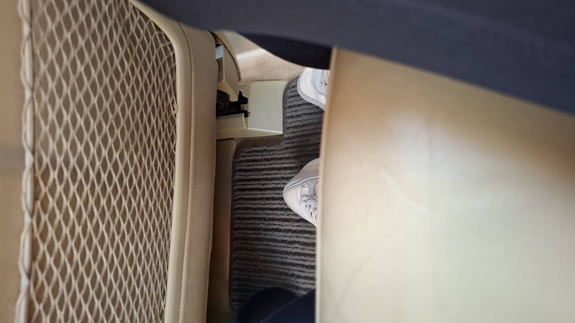 The image shows a person's feet, wearing light-colored sneakers, resting on a grey carpeted floor inside a vehicle. The feet are positioned in the footwell, with a beige mesh pocket visible above. The interior of the vehicle appears to be beige and dark grey. The image was taken in the city of Malo Buchino, Bulgaria.
