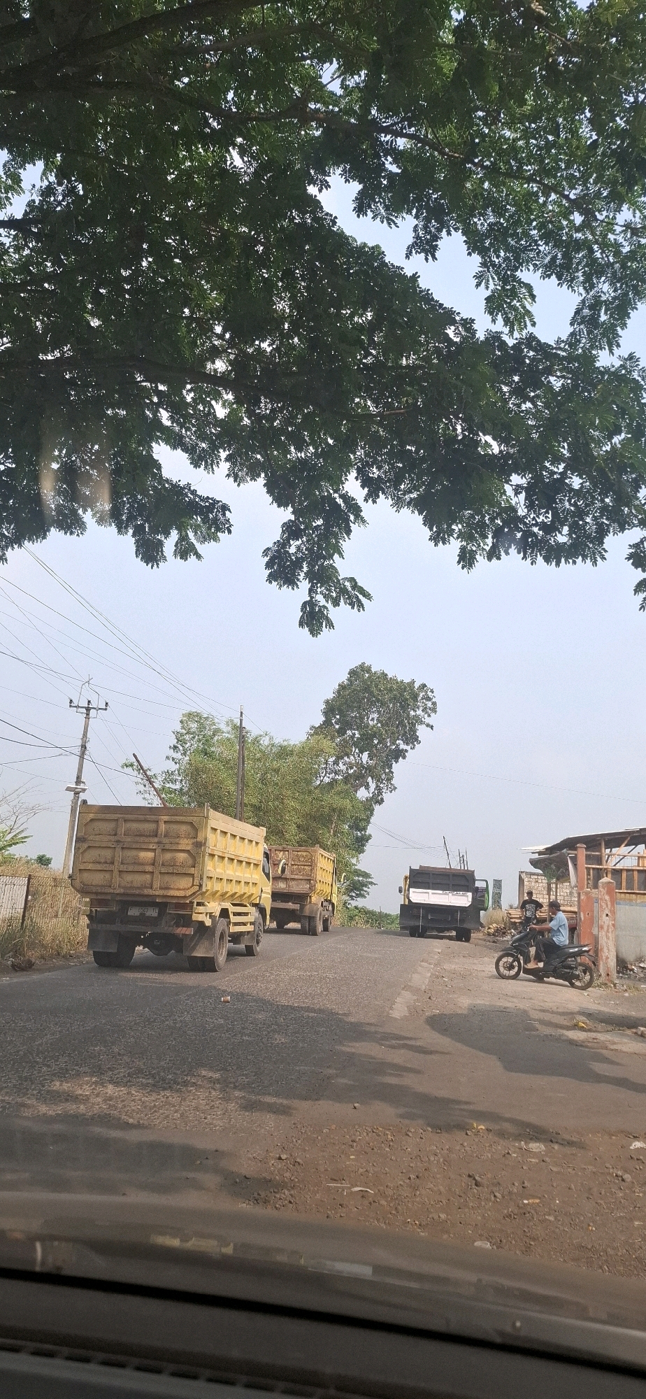 The image shows a sunny day scene on a road in Cibinong Hilir, Indonesia. The foreground is a car's dashboard, suggesting the photo was taken from inside a vehicle. The road is paved with asphalt and dirt, with some loose gravel. In the middle ground, three yellow dump trucks are driving on the road. The first truck is closest to the viewer and is seen from the rear. The second truck is behind the first and is also a dump truck. Further down the road, a third, larger truck with a black bed is visible. To the right of the road, a person is riding a black motorcycle. Behind the motorcycle rider, a building with a corrugated metal roof is partially visible, and two other men are standing near it. The background features utility poles and power lines, with lush green trees framing the top of the image. The sky is clear and bright.