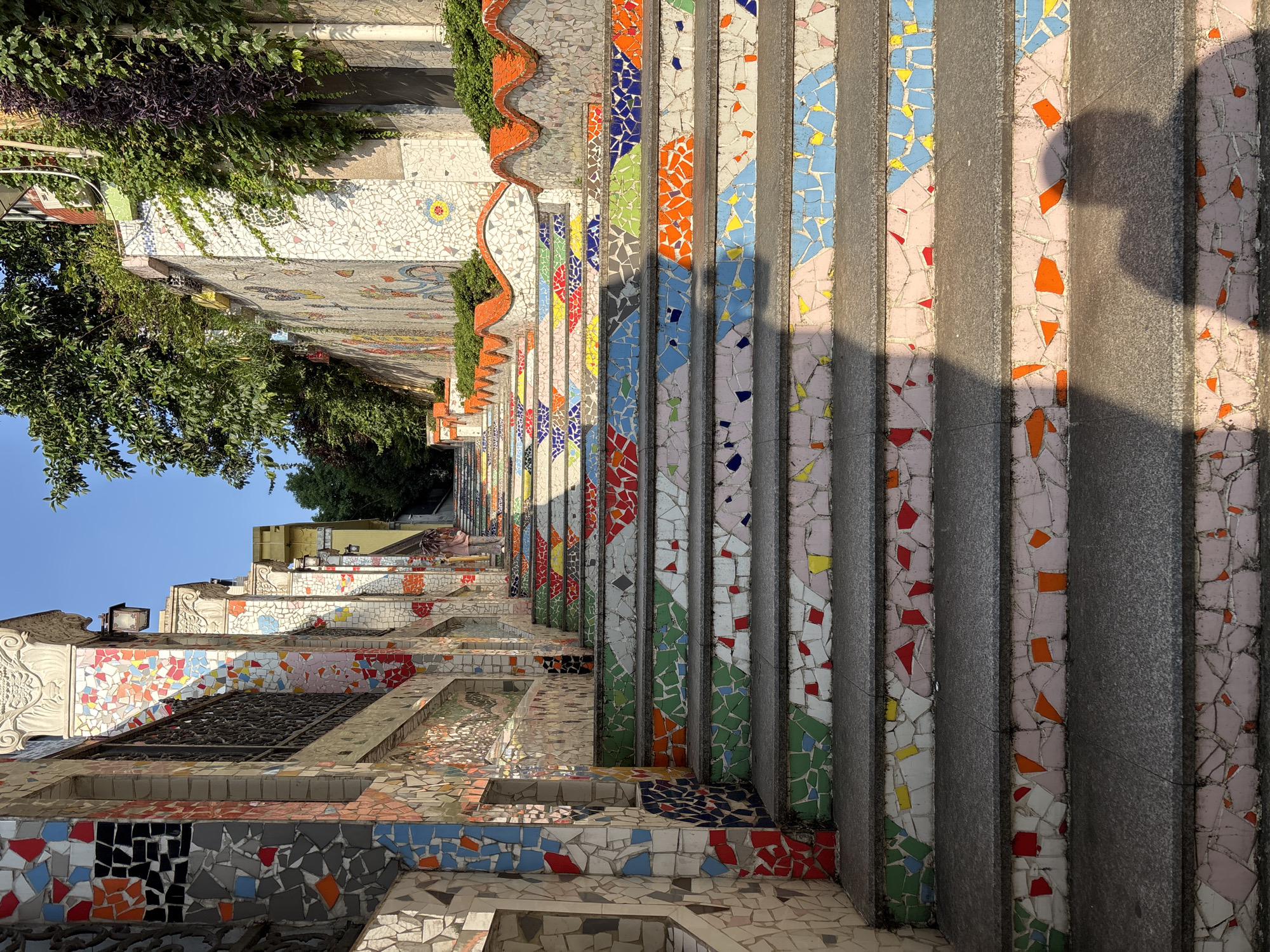 This is a low-angle, full outdoor shot of a vibrantly decorated staircase, leading up a slope in Yuzhong District, China. The steps are adorned with colorful, mosaic tiles in various geometric patterns and fragmented designs. The tiles feature a wide spectrum of colors, including red, orange, yellow, green, blue, pink, and black. The risers of the steps are particularly striking, showcasing a lively arrangement of these mosaic pieces. The sides of the staircase and the adjacent walls are also extensively decorated with similar mosaic work, creating a visually rich and textured environment. Lush green foliage and trees are visible at the top of the staircase and along the side walls, indicating an outdoor setting with vegetation. The sky above is a clear, bright blue. A shadow of a person, likely the photographer, is cast across the lower steps. In the distance, at the top of the staircase, a person is visible, walking away from the camera and carrying an umbrella. The overall scene suggests a public space, possibly a park or a street, designed with artistic embellishments.