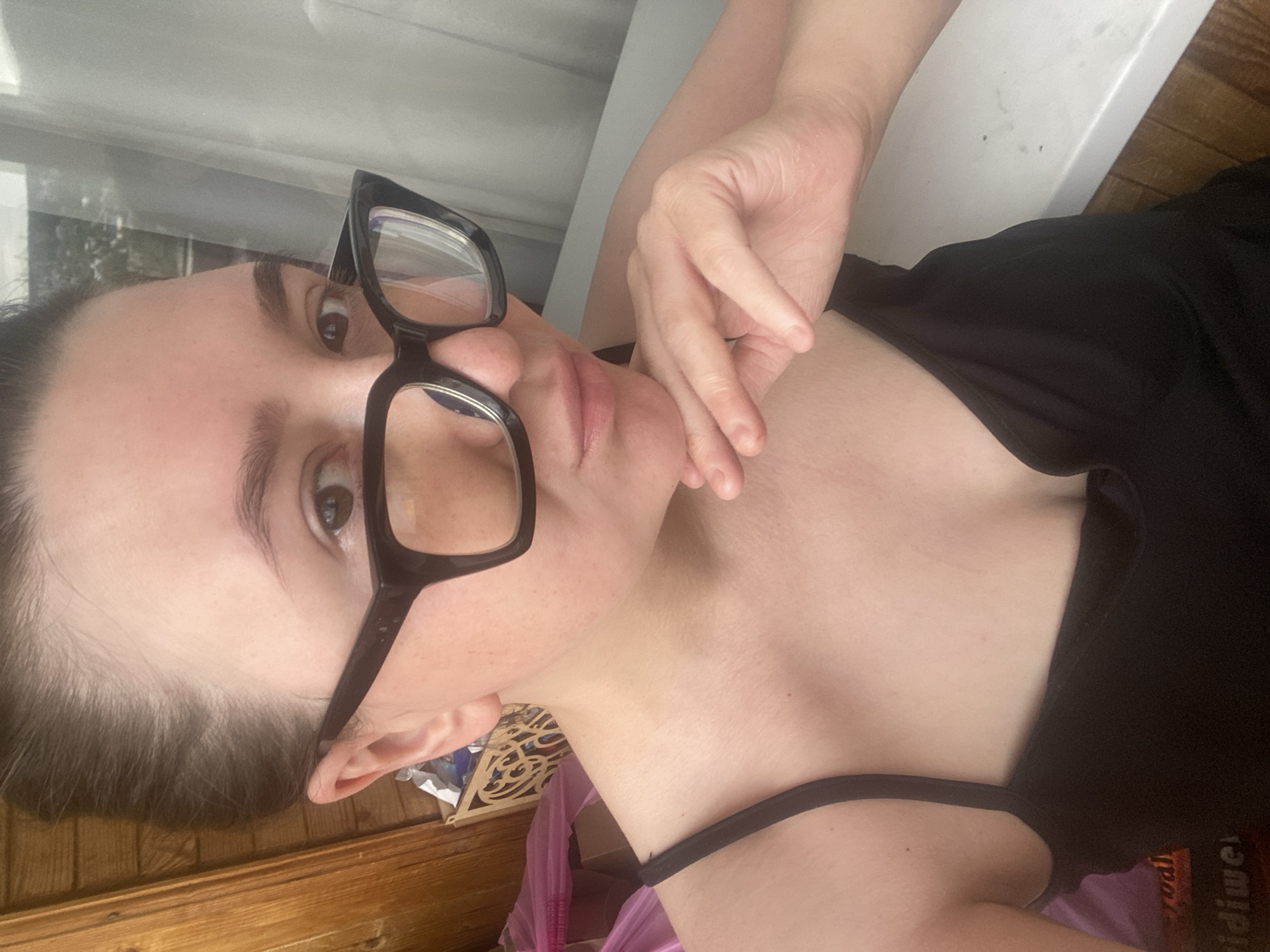 A person with fair skin and dark hair is seen from a close, angled perspective. They are wearing black-rimmed glasses and a black top with thin straps. Their hand is positioned near their face. The background includes a window with natural light and a glimpse of what appears to be interior clutter on a wooden surface. There is no information in the image to confirm a specific city location.
