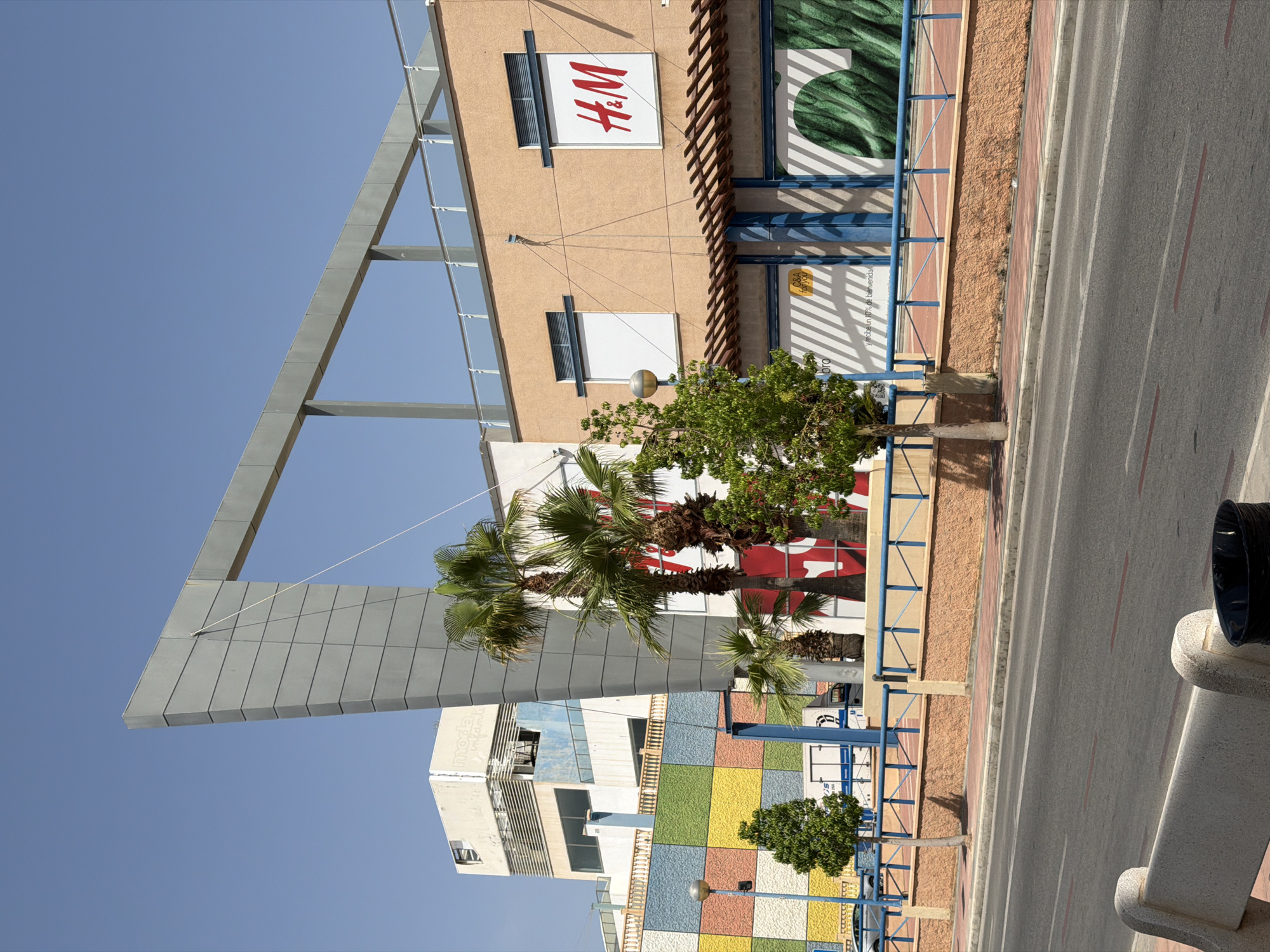 The image shows a street scene in La Cala, Spain, on a clear, sunny day. The foreground features a paved road with red lane markings and a light-colored concrete bench on the left side, next to a black trash can. A sidewalk with red paving stones runs along the edge of the road. Behind the sidewalk, a low, textured orange wall topped with a blue metal railing encloses a raised area with plants and trees.
Further back, a brightly colored building facade with a geometric pattern of squares in yellow, green, blue, and pink is visible. Adjacent to this is a modern building with a large, angular gray structure extending upwards, contrasting with the blue sky. Palm trees are scattered throughout the scene, adding to the Mediterranean feel.
On the right side of the image, a tan-colored building features a prominent H&M logo in red and white, indicating a retail establishment. Windows with horizontal blinds are visible above the storefront. Below the H&M sign, a display window shows a green, textured image. Another storefront to the left displays a "C&A for you" sign. In the background, a taller, lighter-colored building with visible windows and some signs appears partially dilapidated. The overall scene depicts a commercial area with a mix of modern and older architecture under a bright sky.