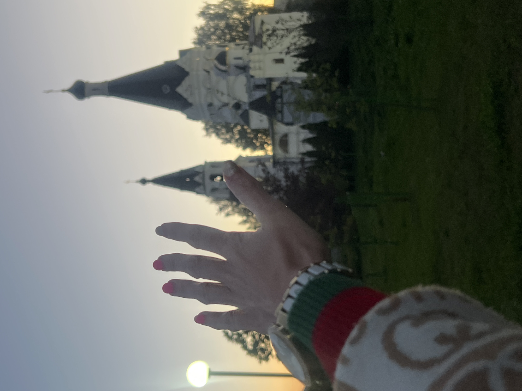 The image, taken outdoors in Krasnoe-na-Volge, Russia, features a person's outstretched hand in the foreground, with its palm facing the viewer. The hand has bright pink nail polish on its fingernails. The person is wearing a watch with a silver band and a bracelet with green and red stripes. Part of their clothing is visible, featuring a light-colored fabric with a pattern of beige interlocking shapes.
In the background, two white buildings with dark, pointed roofs and spires are prominent. These structures appear to be churches, likely religious buildings common in Russian landscapes. They are partially obscured by lush green trees and foliage. The sky suggests it is either early morning or late evening, with soft, diffused light indicating either sunrise or sunset. The overall lighting is warm and hazy, creating a serene atmosphere. A streetlamp with a glowing light is visible in the lower portion of the image, adding to the sense of an outdoor setting at dusk or dawn. The orientation of the image is sideways.