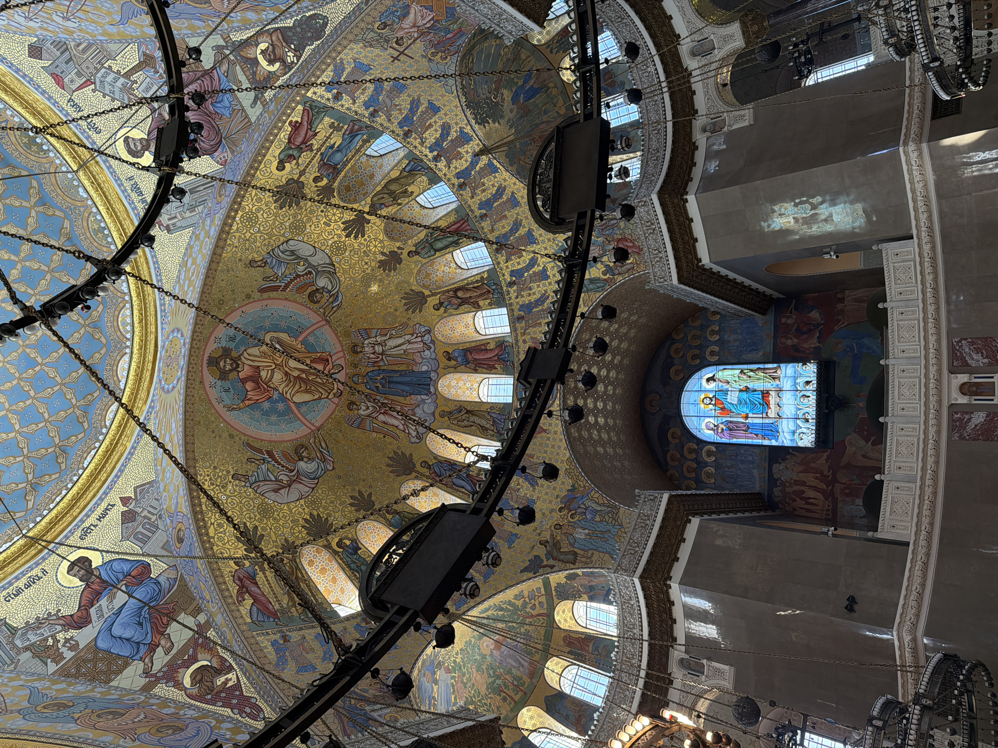The image shows the interior of a church, specifically looking upwards at the dome. The dome is adorned with Byzantine-style mosaics depicting religious figures and scenes. Several arched windows punctuate the dome. A large, elaborate chandelier hangs suspended from the dome, partially obscuring the artwork. The walls are decorated with mosaics and paintings. A stained glass window is visible on one side. The location is likely St. Nicholas Naval Cathedral in Kronstadt, Russia, given the style of architecture and decoration.