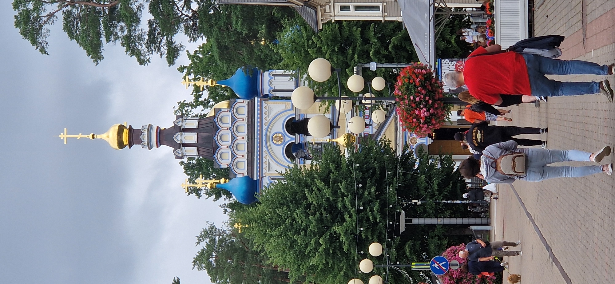 The image depicts a street scene in front of an Eastern Orthodox church, characterized by its blue domes and ornate architectural details. In the foreground, several people are walking away from the camera on a cobblestone street. A woman in the immediate foreground wears a grey hooded sweatshirt and light blue jeans, carrying a small backpack. To her right, a man in a red t-shirt and blue jeans walks with a dark jacket slung over his shoulder. Further back, a man in a black jacket with white lettering on the back is walking, followed by individuals in orange and black clothing. A small dog on a leash is visible on the left side of the image, being walked by a man in a blue polo shirt and grey pants. The street is lined with trees and lampposts adorned with hanging flower baskets. In the background, the church is the prominent structure, with its golden and blue onion domes reaching towards a cloudy sky. The overall scene suggests a public space, possibly a town square or a pedestrian walkway, during the daytime.