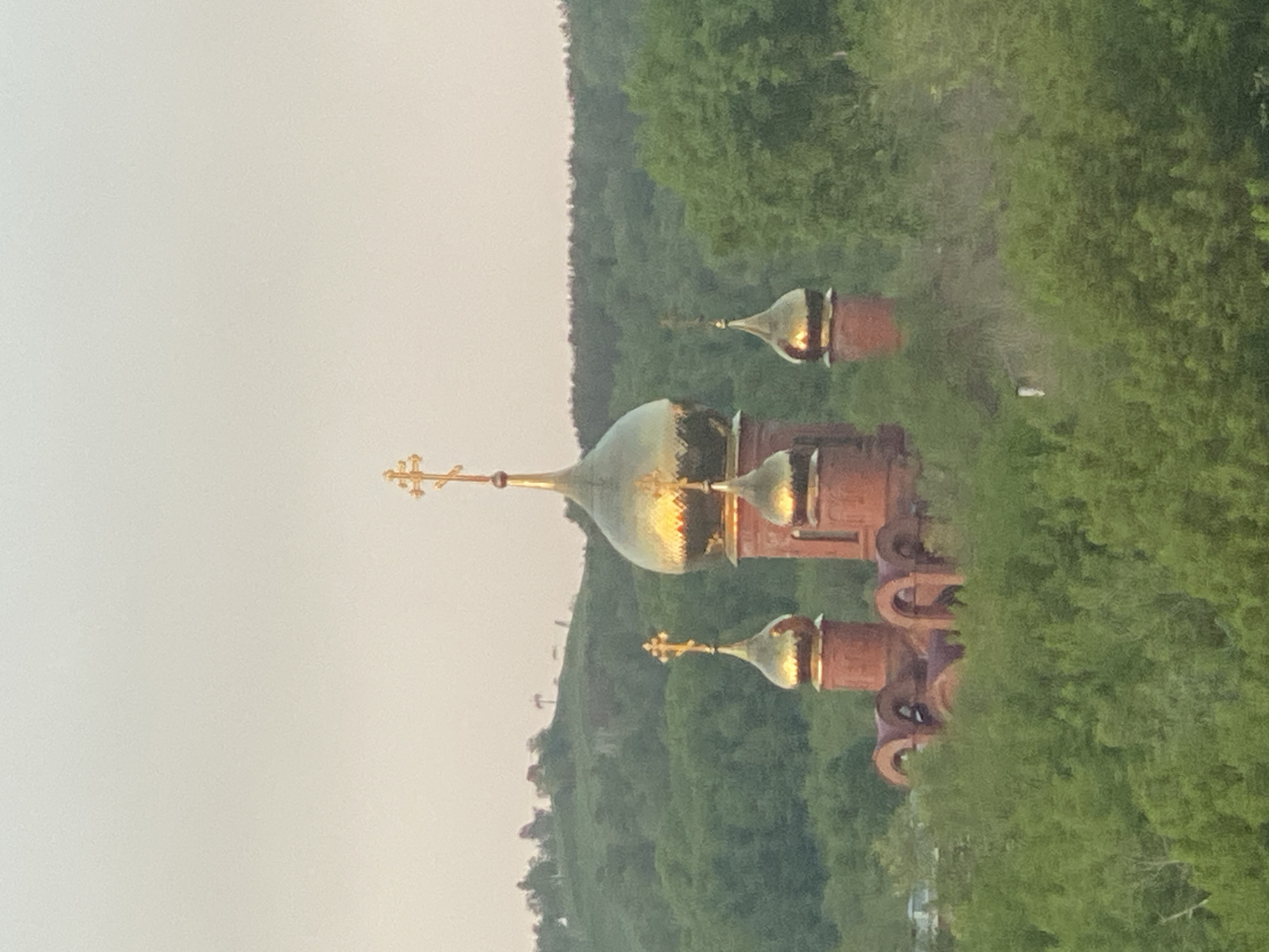 The image depicts a Russian Orthodox church with multiple golden domes and crosses atop, set against a backdrop of a green, forested hillside. The sky is overcast. The church appears to be constructed of red brick. The location is likely in or near Novokuznetsk, Russia, as the domes are representative of this style of architecture.