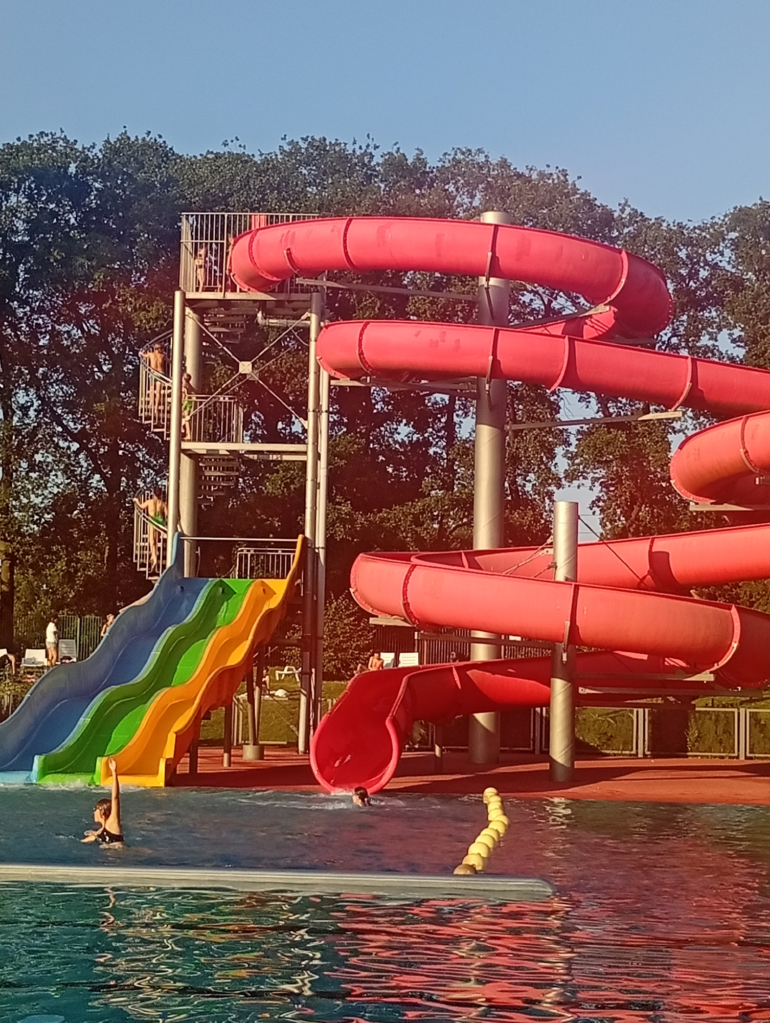 The image shows an outdoor water park with several large red water slides and a multi-colored slide. The water park is surrounded by trees and a clear blue sky. There are several people visible in the image, some are on the slides or climbing stairs, while others are in the pool. The water in the pool reflects the red slides and the surrounding trees. The scene appears to be a sunny day at a recreational facility. The location is likely a water park in Wrocław, Poland, given the context provided by the user.